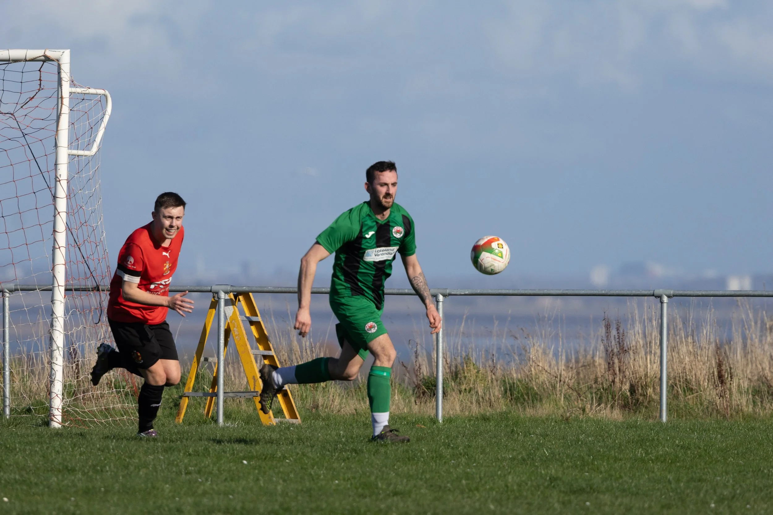 Two soccer players, one in a red jersey and one in a green jersey, are on a grass field near a goalpost during a match. The player in green is chasing the ball, which is in mid-air, while the player in red is running behind him.