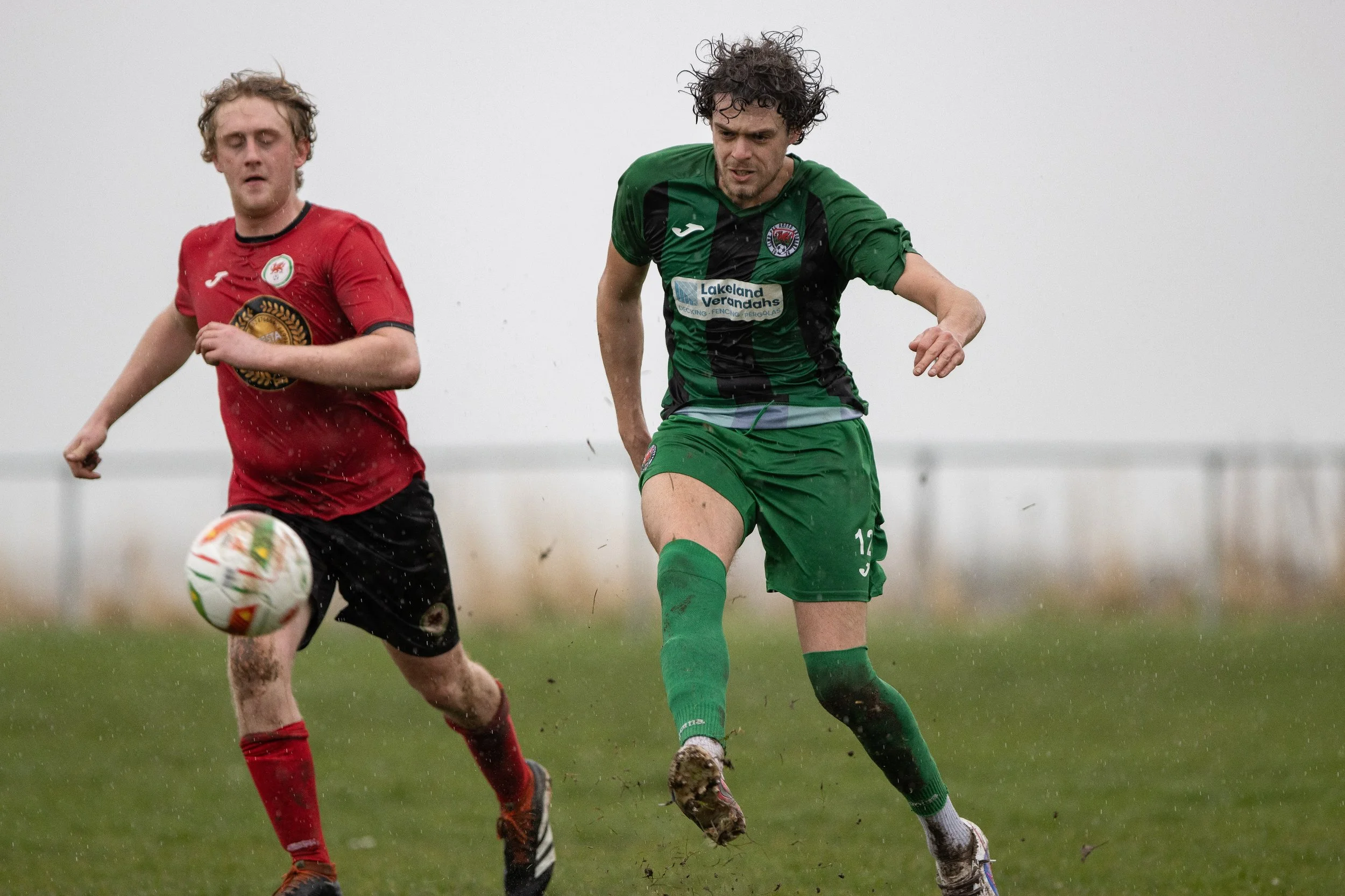 Two soccer players running on a muddy field during a game in rainy weather. One player in a red jersey and black shorts, the other in green jersey and shorts. The green-clad player is focused on the ball, which is in front of the red player.