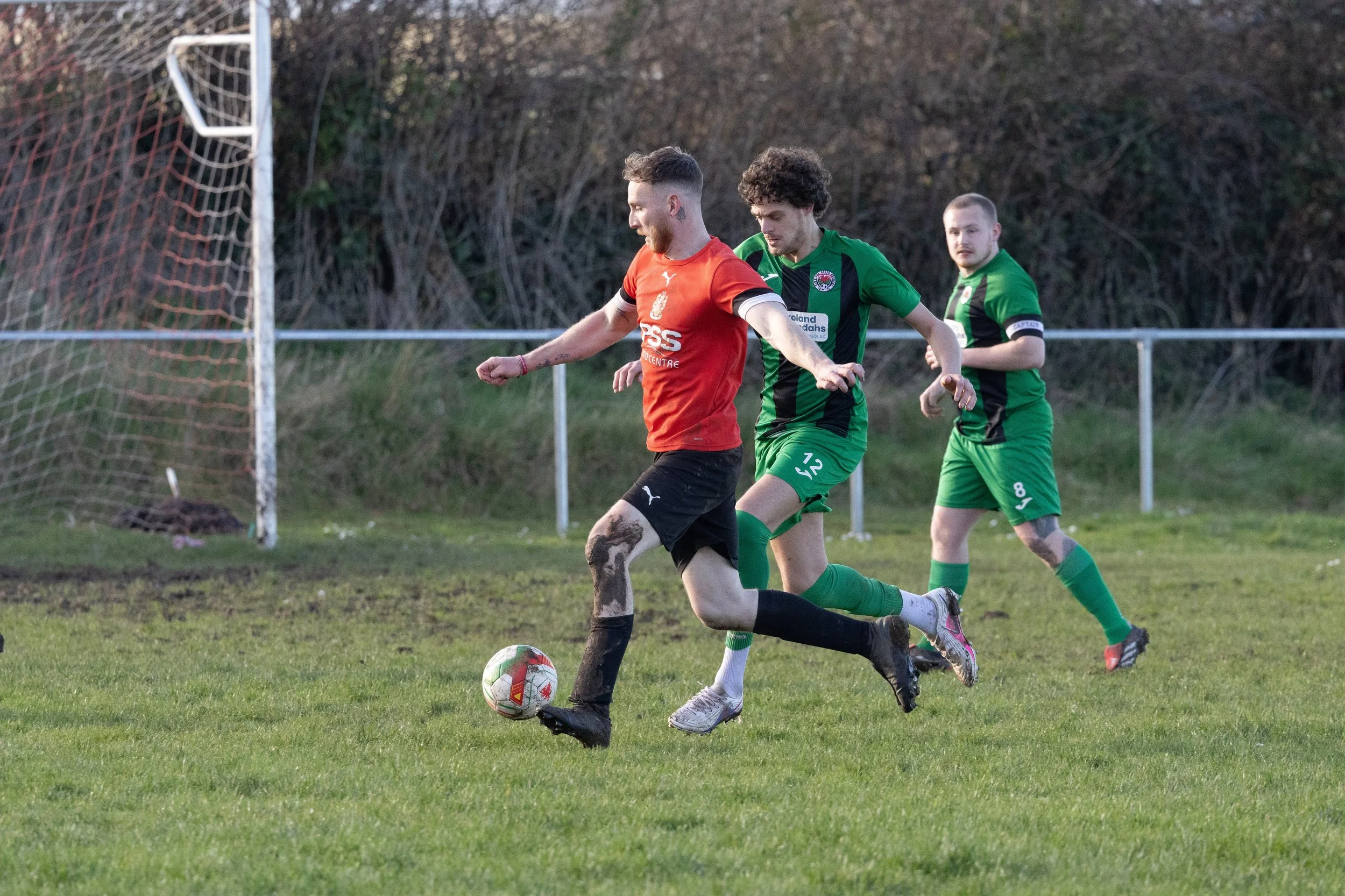 Three soccer players chasing after a ball on a grassy field, with one player wearing an orange jersey and black shorts, and two players wearing green jerseys and shorts, near a soccer goal with a net, in a rural outdoor setting.