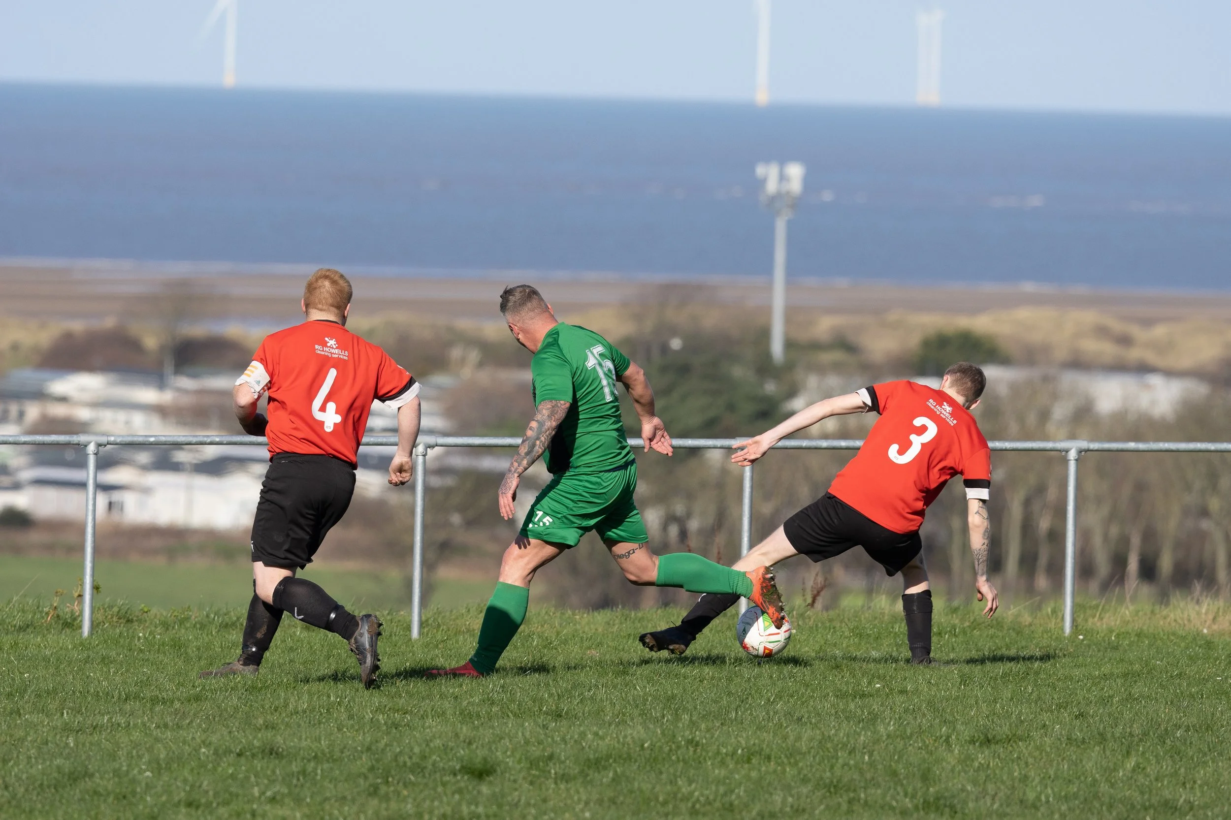 Three soccer players on a field near a fence, with a large body of water and wind turbines in the background. Two players are wearing red jerseys with numbers 4 and 3, and one player is wearing a green jersey with number 15, actively competing for th