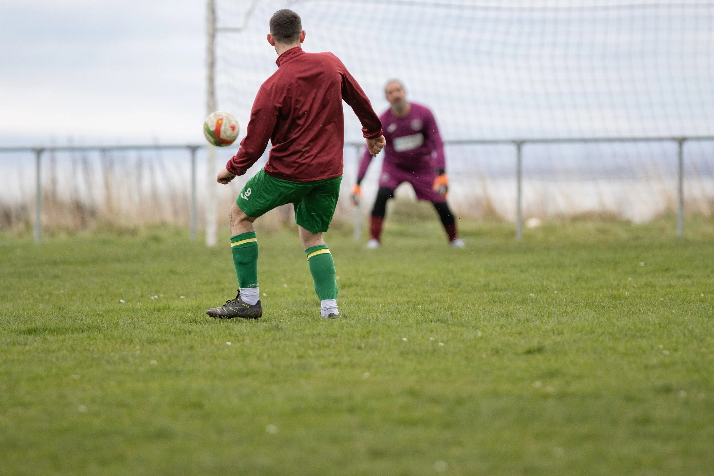 A soccer player in a red jacket and green shorts kicking the ball towards the goal with a goalkeeper in a purple uniform preparing to block the shot.