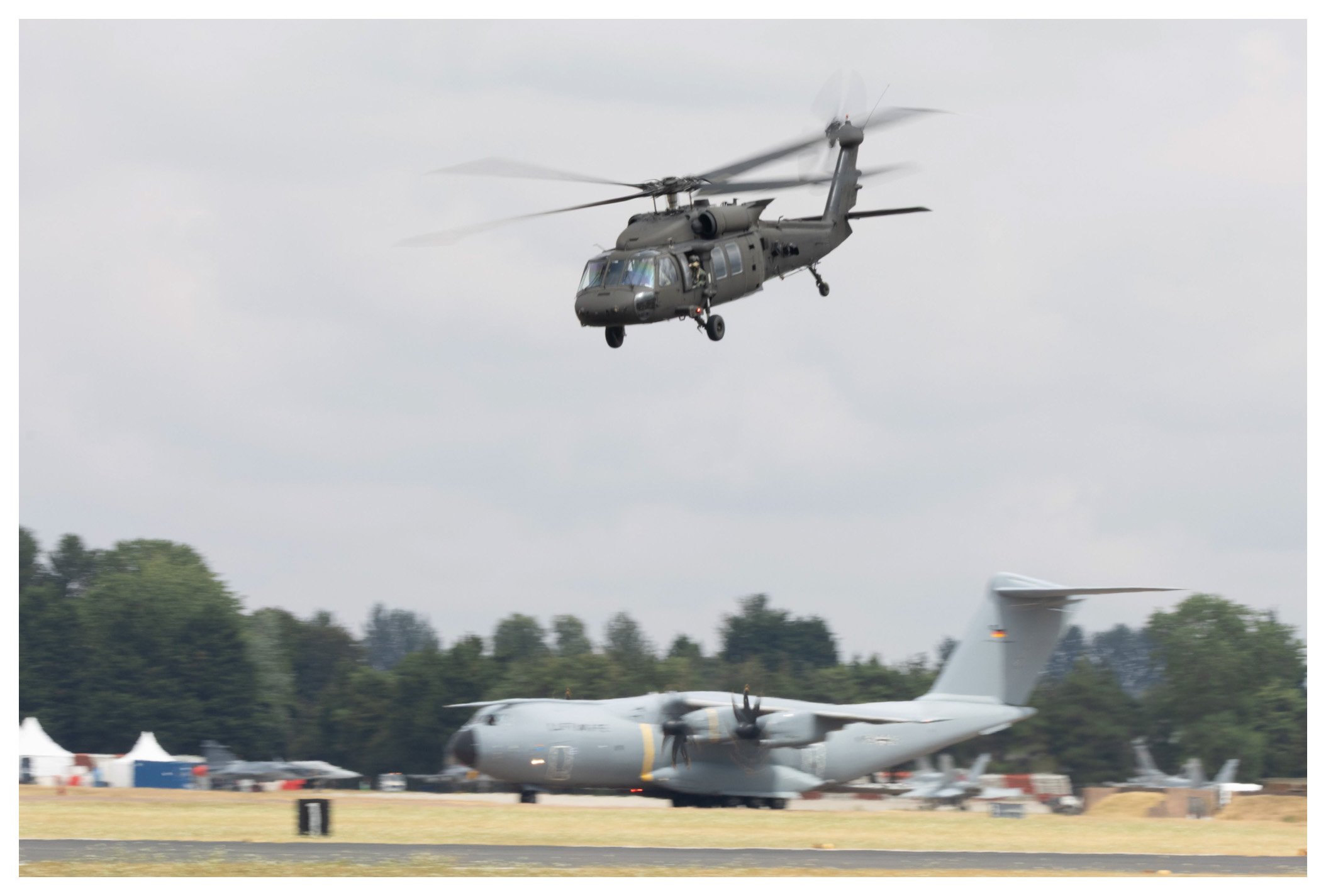 A military helicopter flying above a large cargo plane parked on a tarmac with trees in the background.