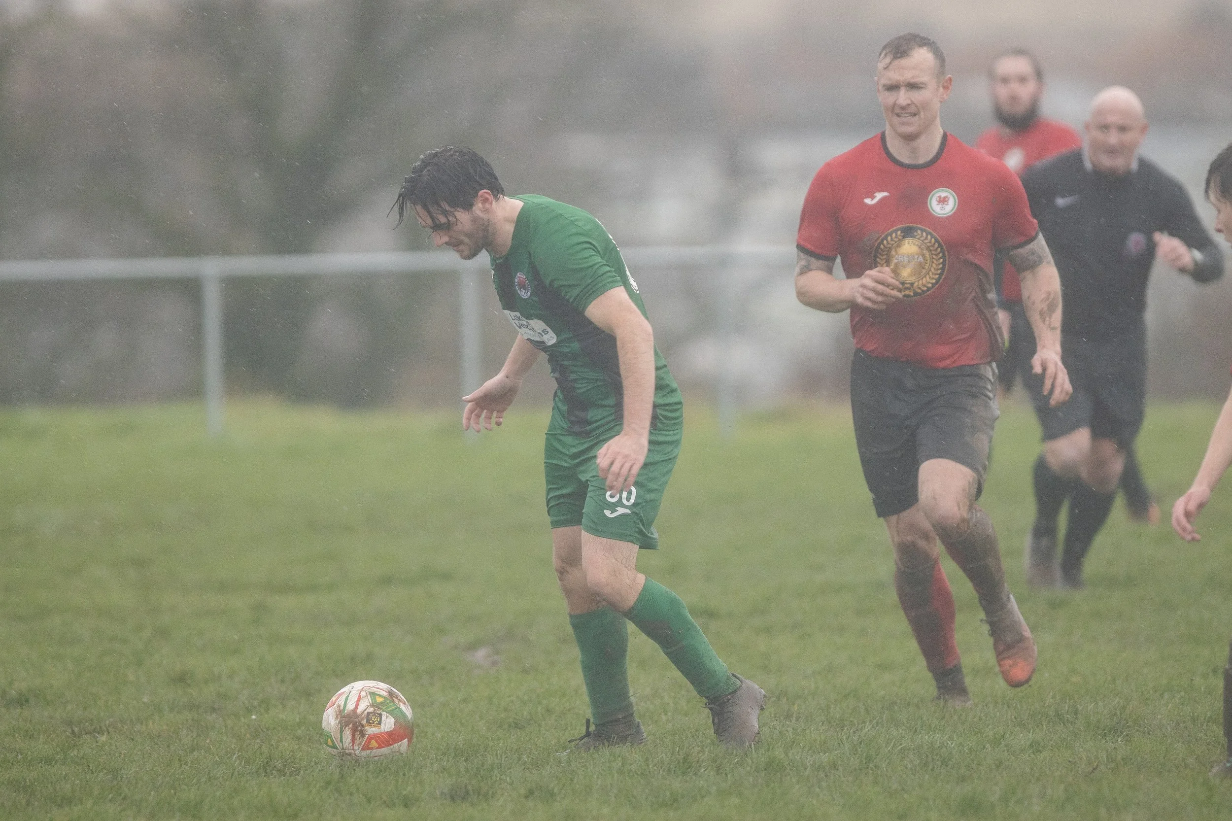 Soccer players playing in the rain on a grass field, with one player in a green uniform looking down at the ball and a player in a red uniform running behind him.