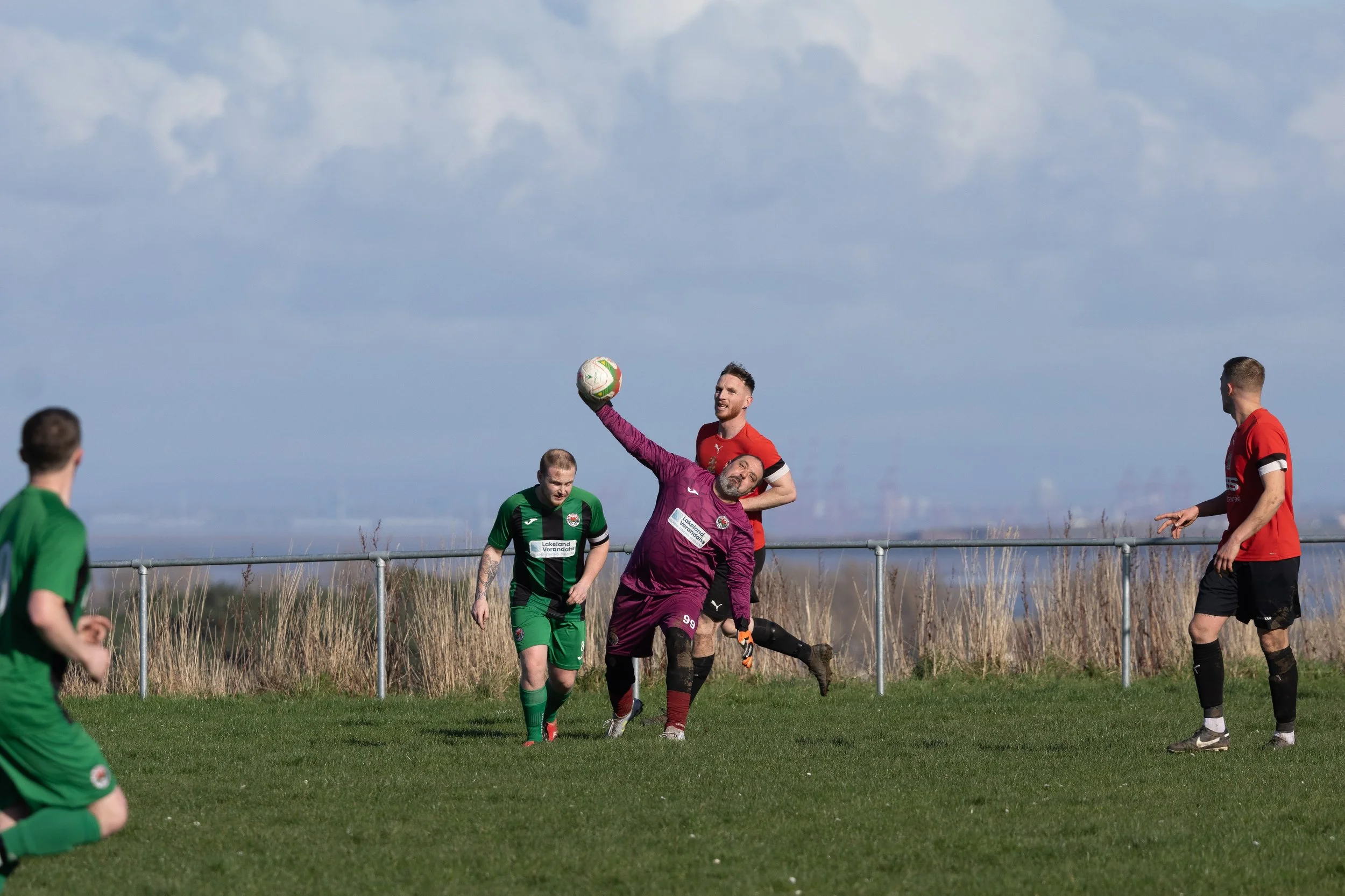 Soccer players on a field during a match, with one player in a purple uniform catching or punching the ball, and other players in red and green uniforms watching or preparing.