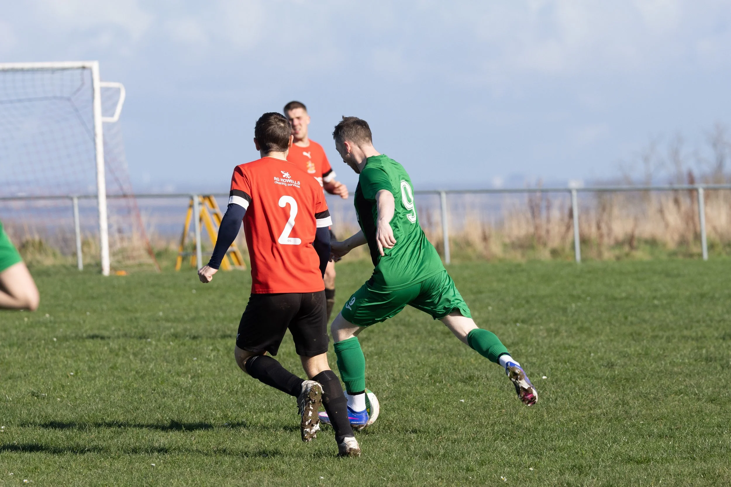 Soccer players in green and red uniforms competing for the ball on a grassy field with goal in background.