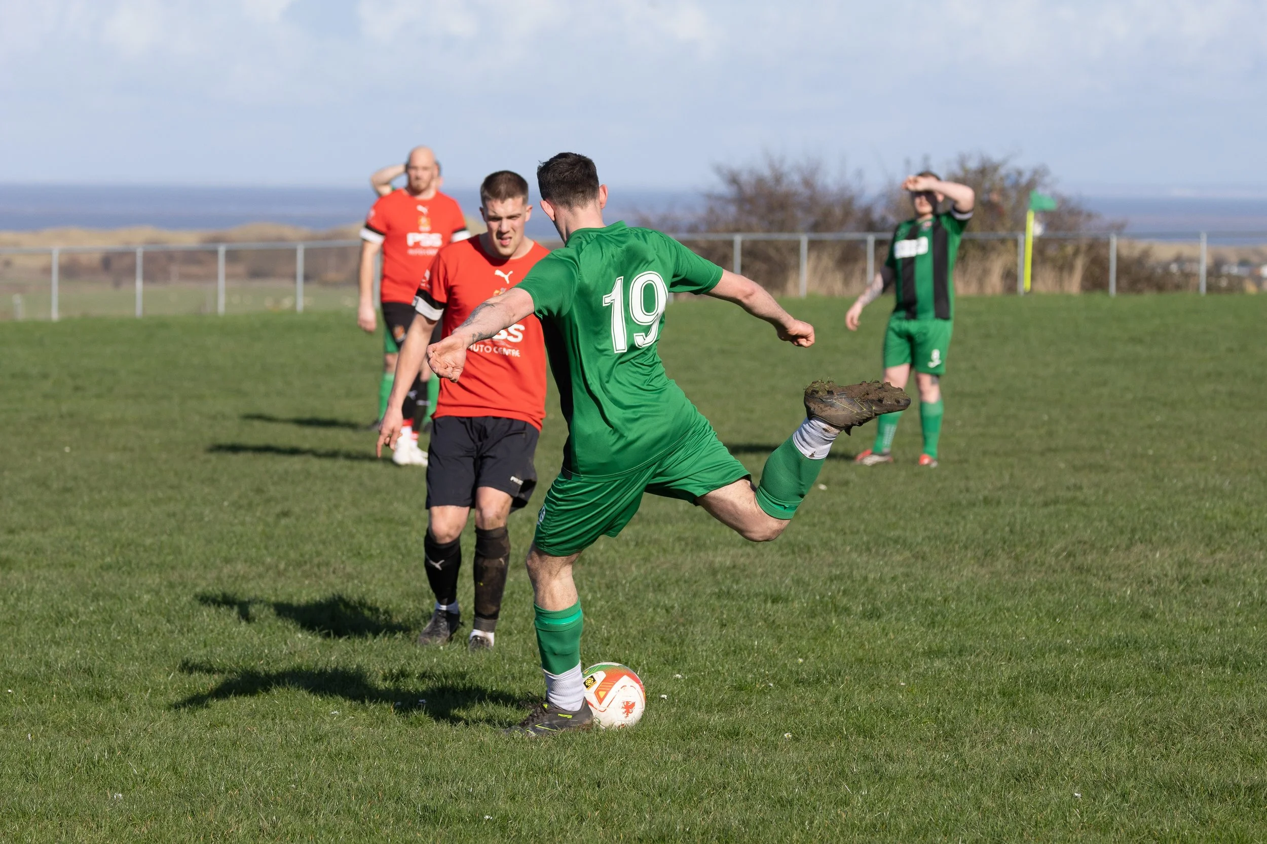 A soccer player in a green uniform with number 19 kicks a ball on a grassy field while several players in red and black uniforms watch, with two additional players in green in the background.
