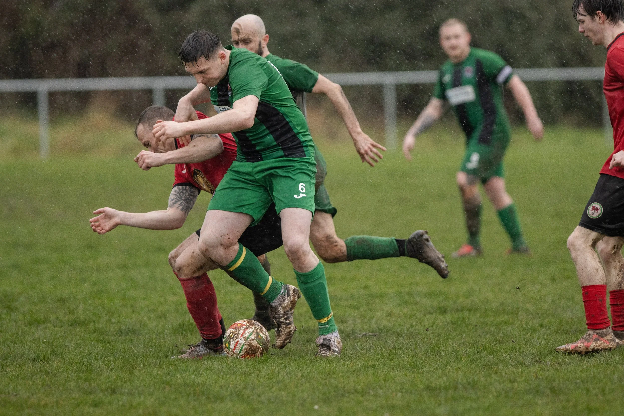 Soccer players fighting for the ball on a muddy field in the rain, with some players wearing green and red uniforms.