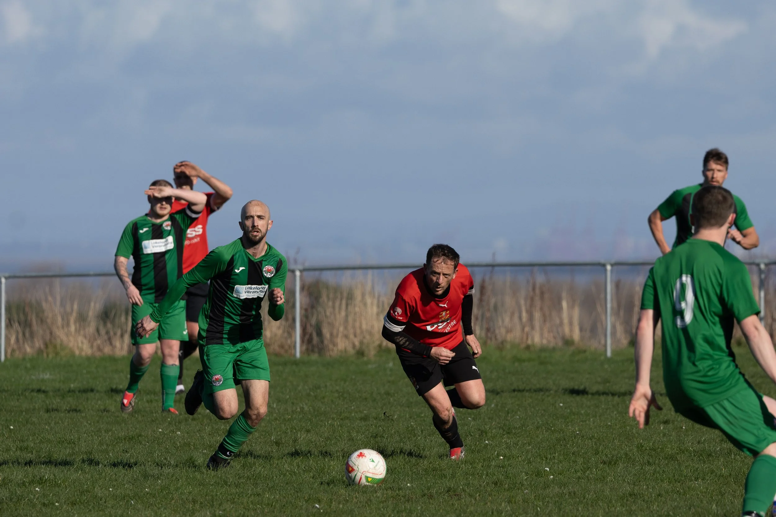 Soccer players on the field, with one player in a red jersey about to kick the ball, while others in green jerseys run or watch during a match