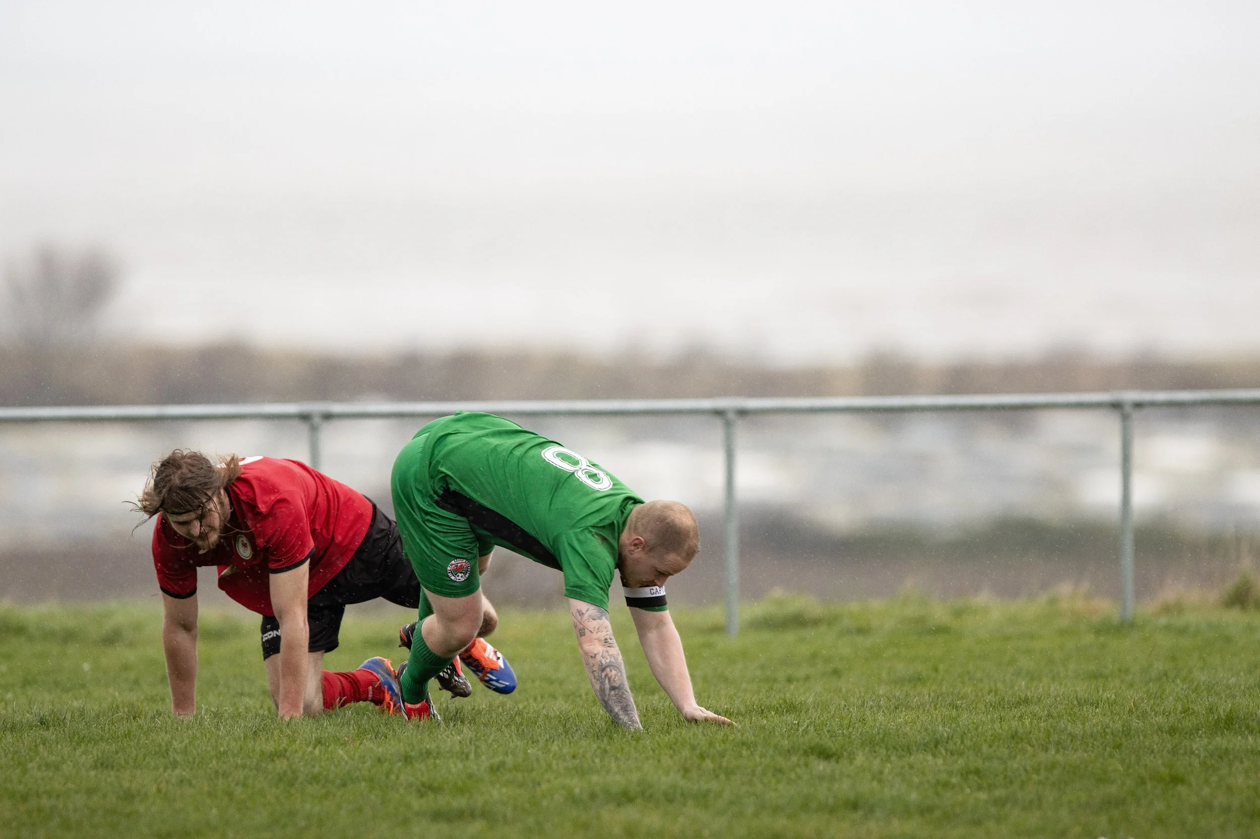 Two rugby players on the ground, one in a red jersey and the other in a green jersey, during a match on a grassy field with a cloudy sky in the background.