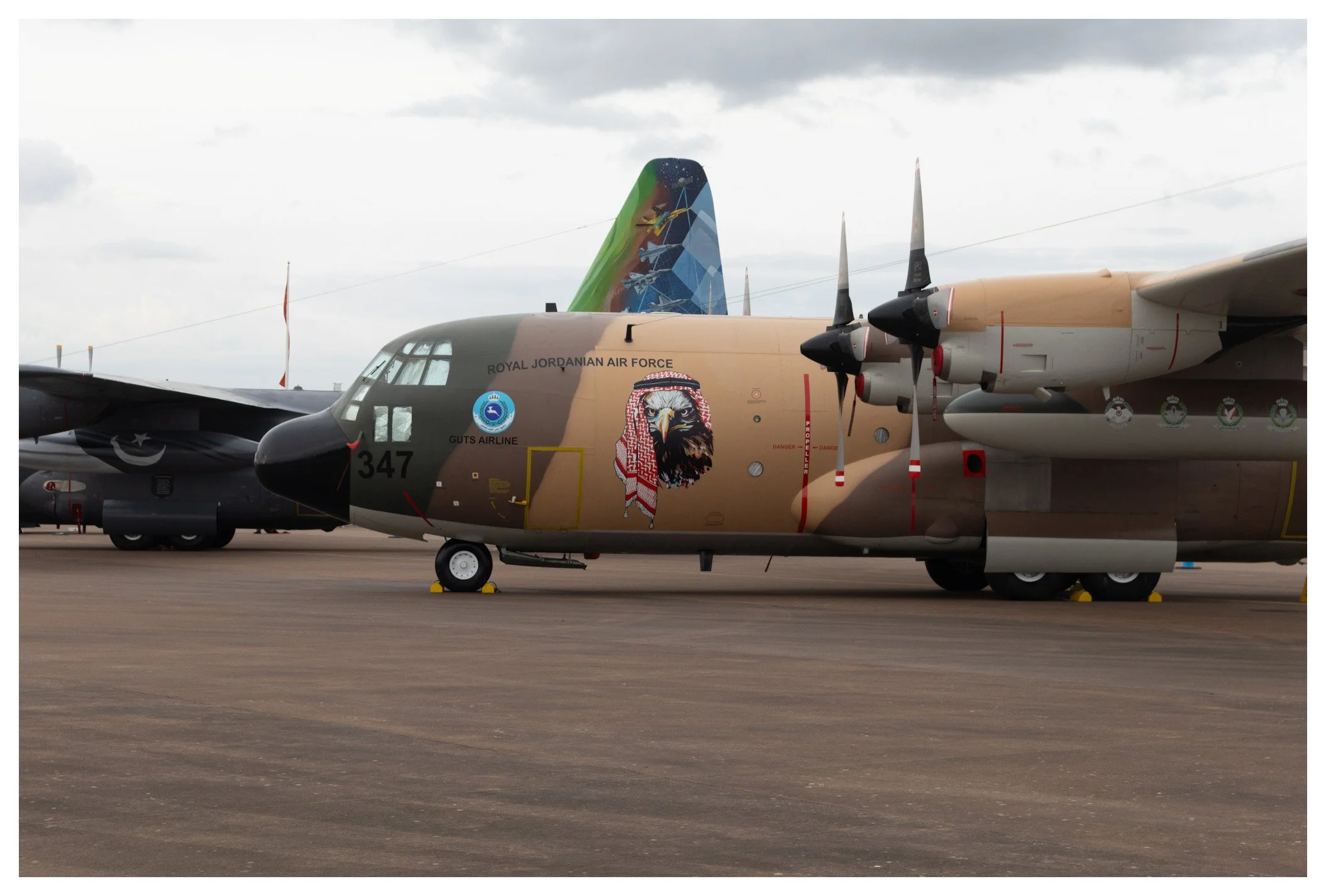 Military aircraft on tarmac with desert camouflage and bold eagle emblem, marked 'Royal Jordanian Air Force' and 'Guts Airline', with a colorful tail featuring a world map design, under cloudy sky.