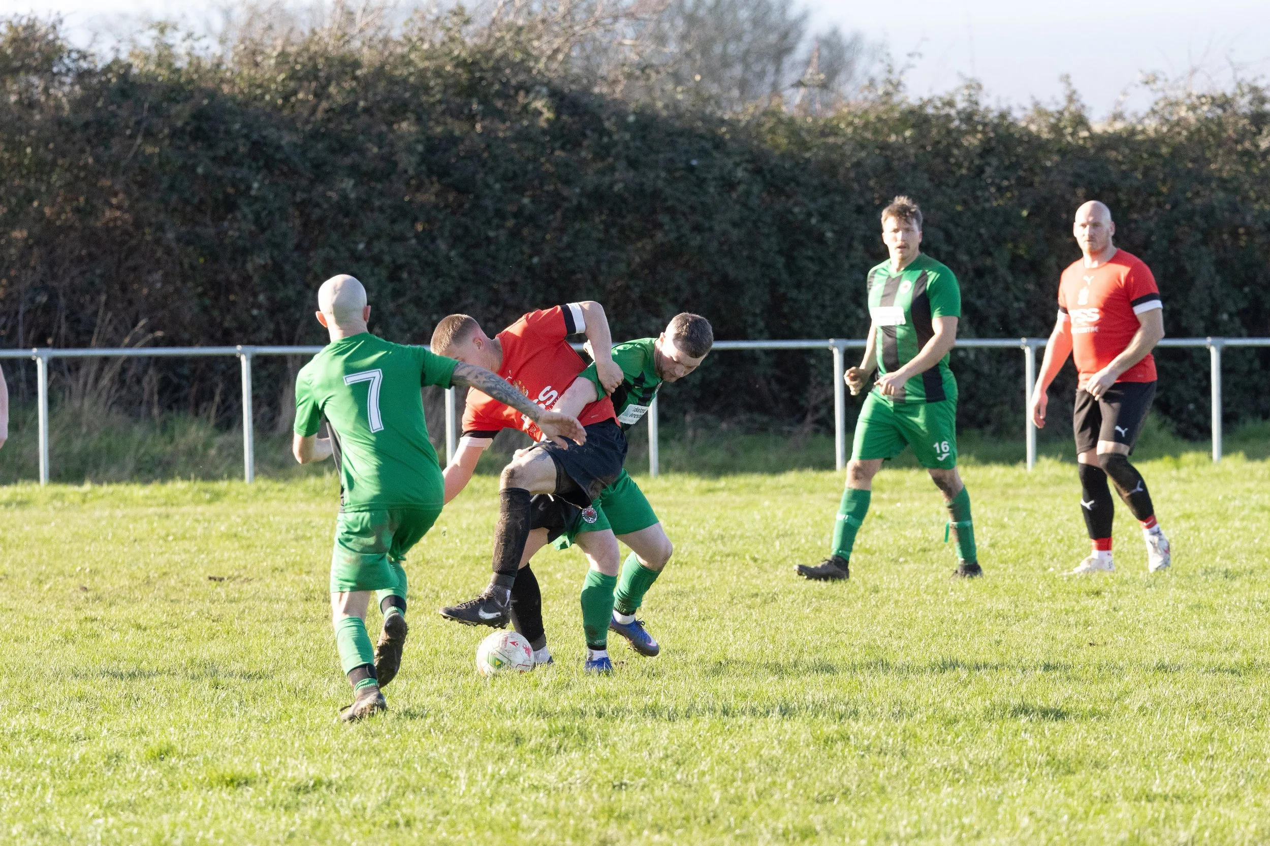 Soccer match with players in green and red uniforms battling for ball on grassy field, with three players observing.