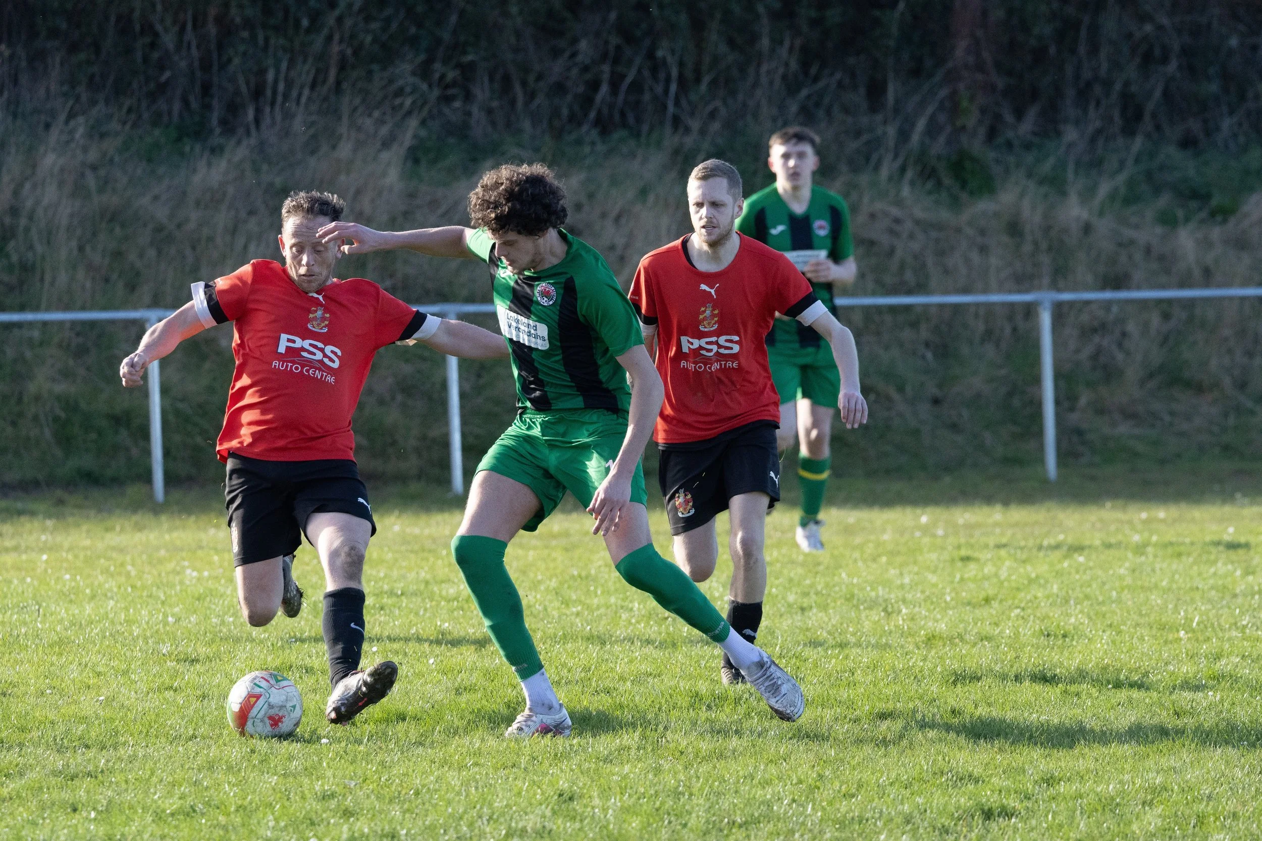 Soccer match with players in green and red jerseys competing for the ball on a grassy field, with a natural background and spectators or officials in the distance.