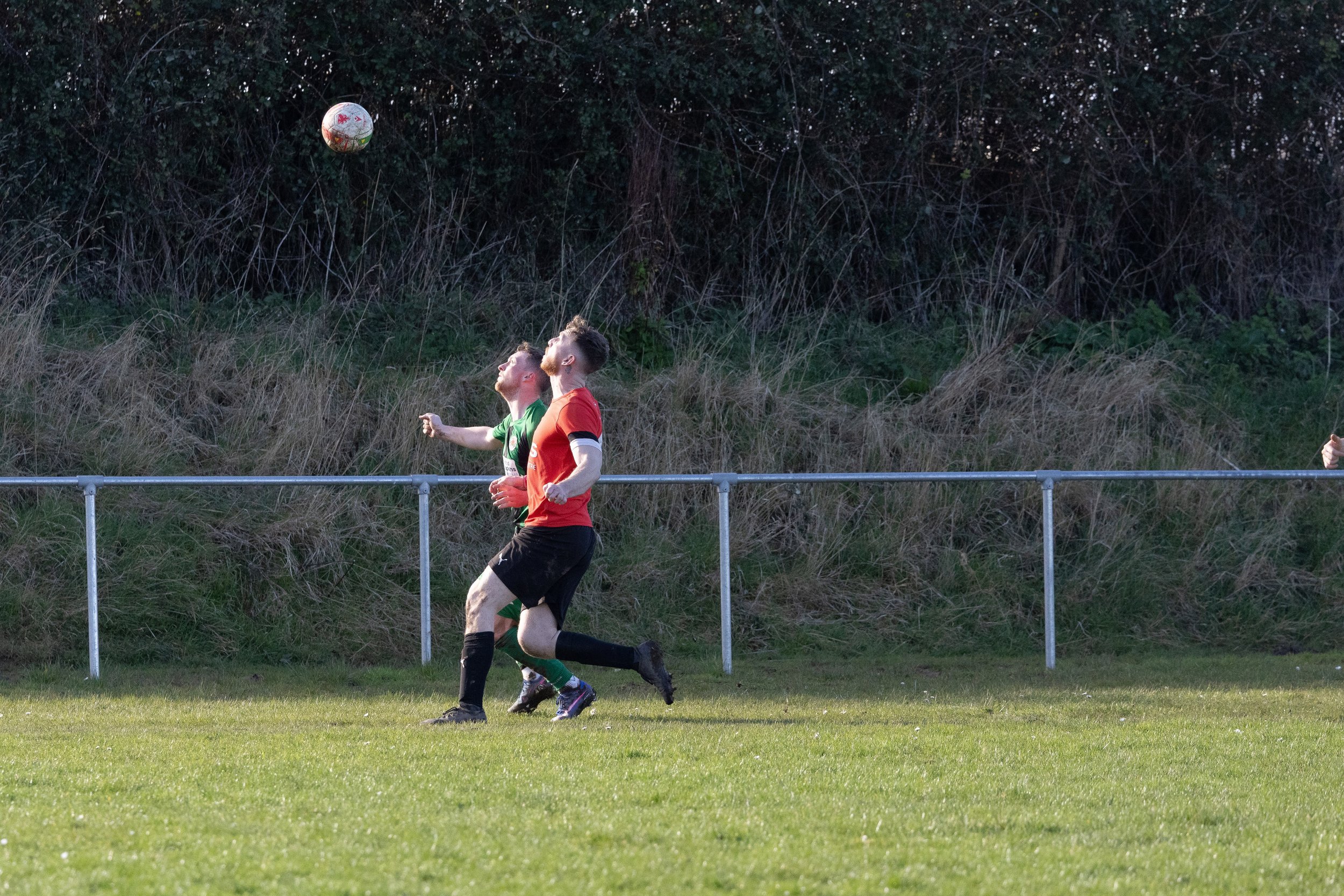 Two soccer players competing for the ball on a grassy field, with a grassy hill and trees in the background.