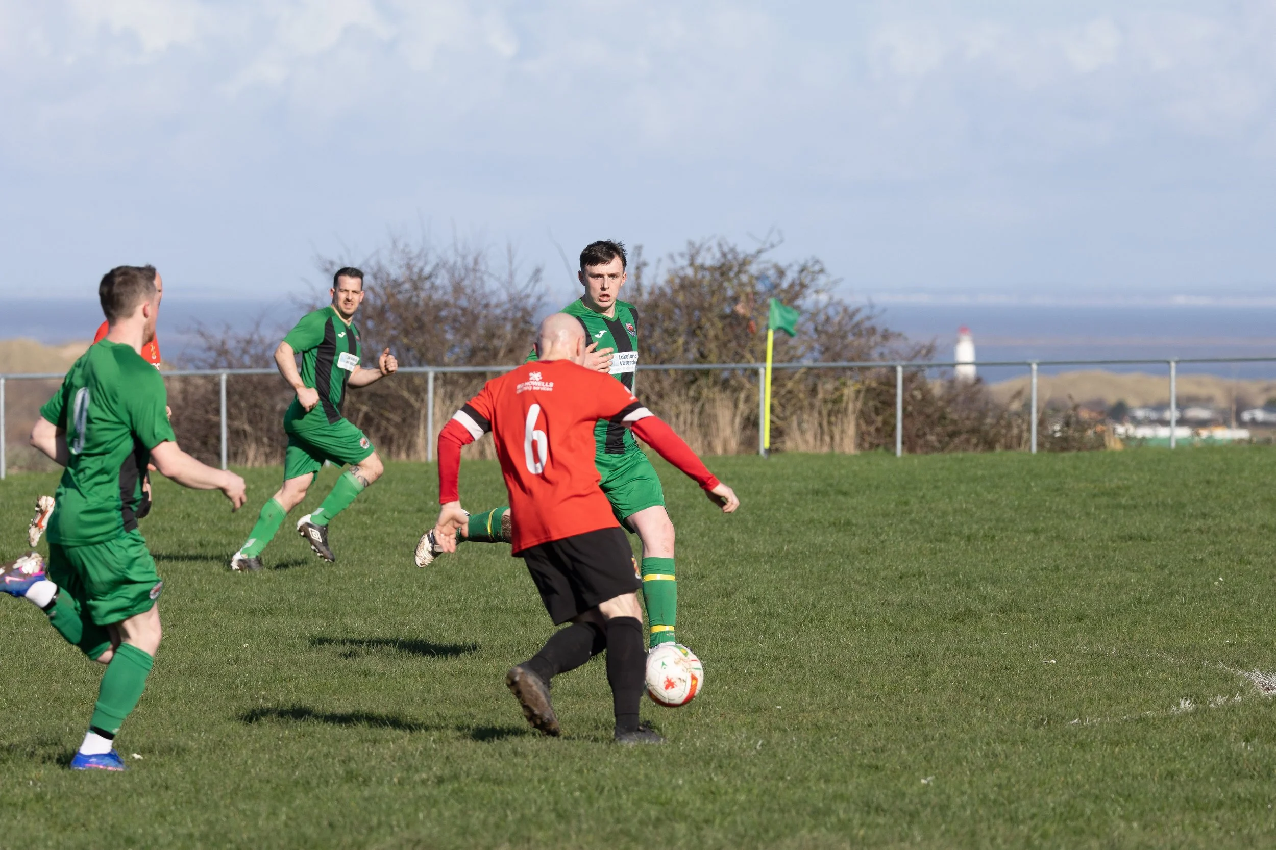 Soccer match with players in green and red jerseys on a grassy field near a fence and lighthouse in the background.