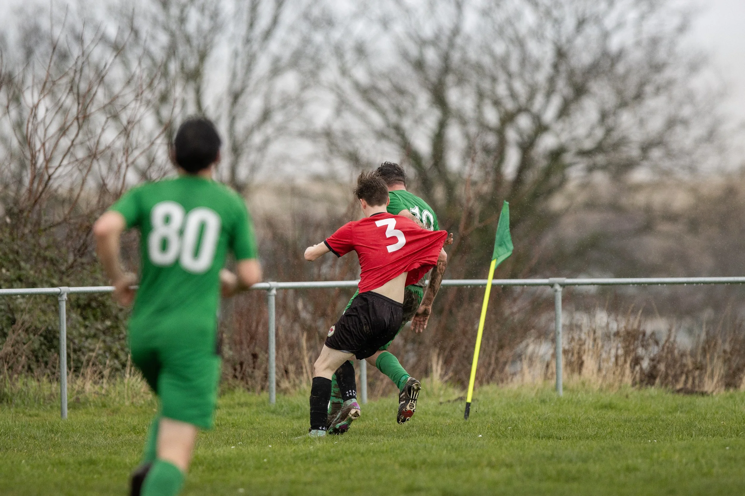 A soccer game where a player in red with the number 3 is kicking a player in green with the number 10, near a corner flag, with another player in green with number 80 watching.