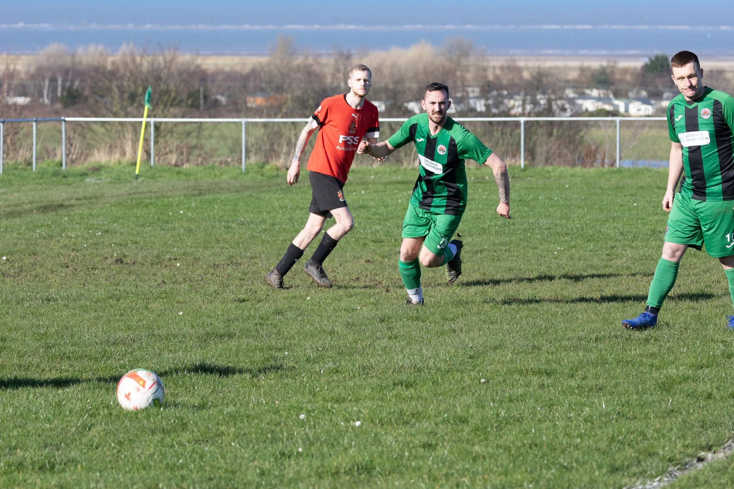 Three soccer players on a green field, two in green and black uniforms and one in red and black, near a white soccer ball with an orange star pattern, during daytime with a distant landscape and cloudy sky.