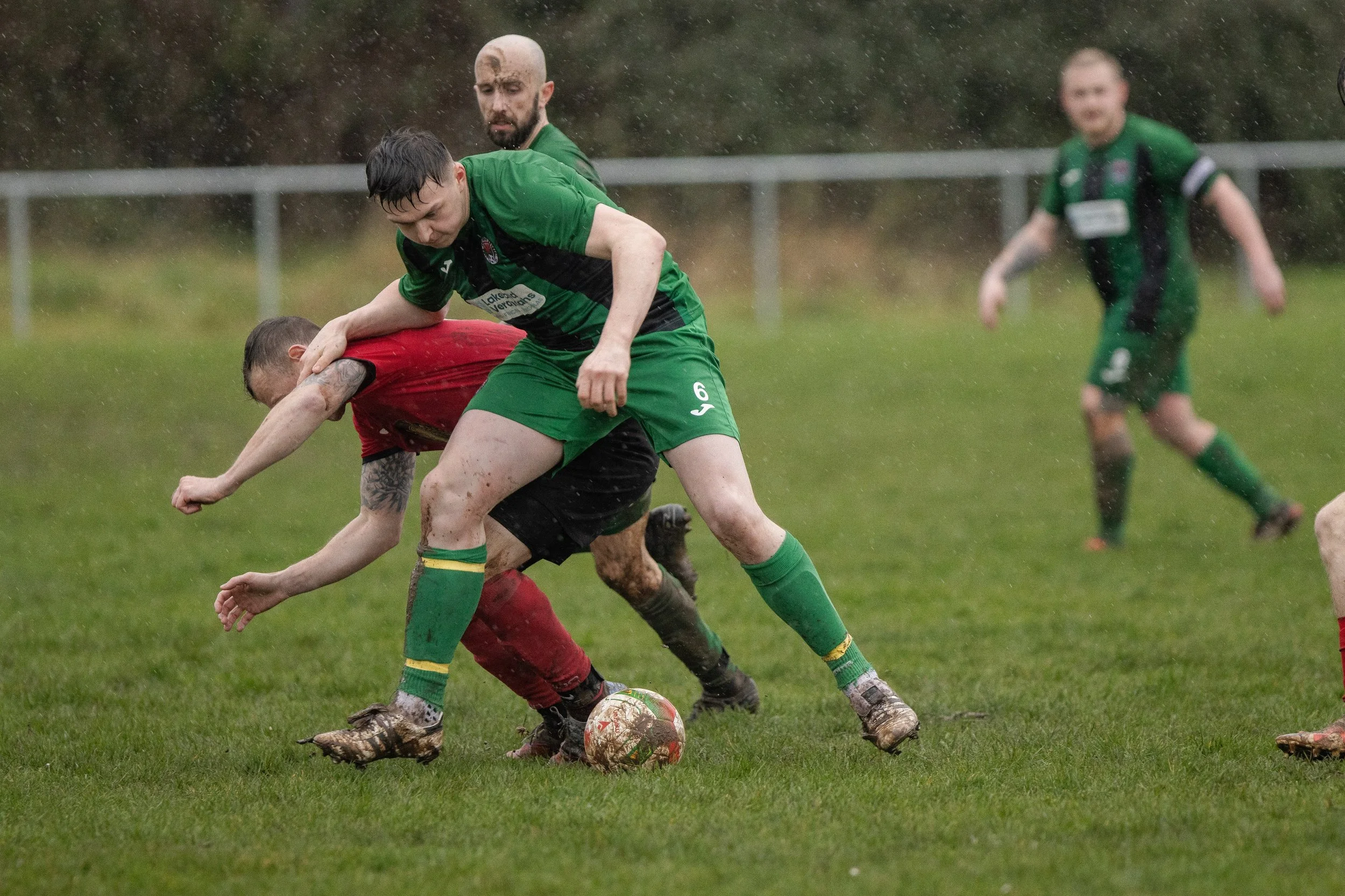 Two soccer players, one in a red jersey and one in a green jersey, compete for the ball during a match in rainy weather, while a third player in green watches nearby.