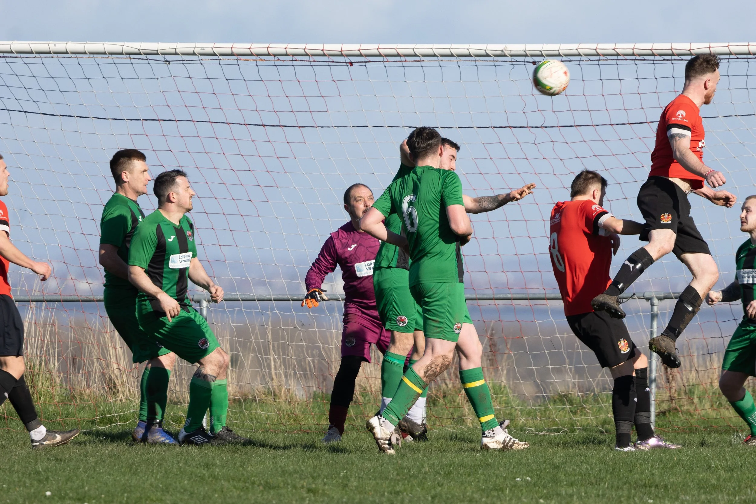 Soccer players jumping to head the ball near the goal with three players in green and two players in red, a goalkeeper in purple, and a net in the background.