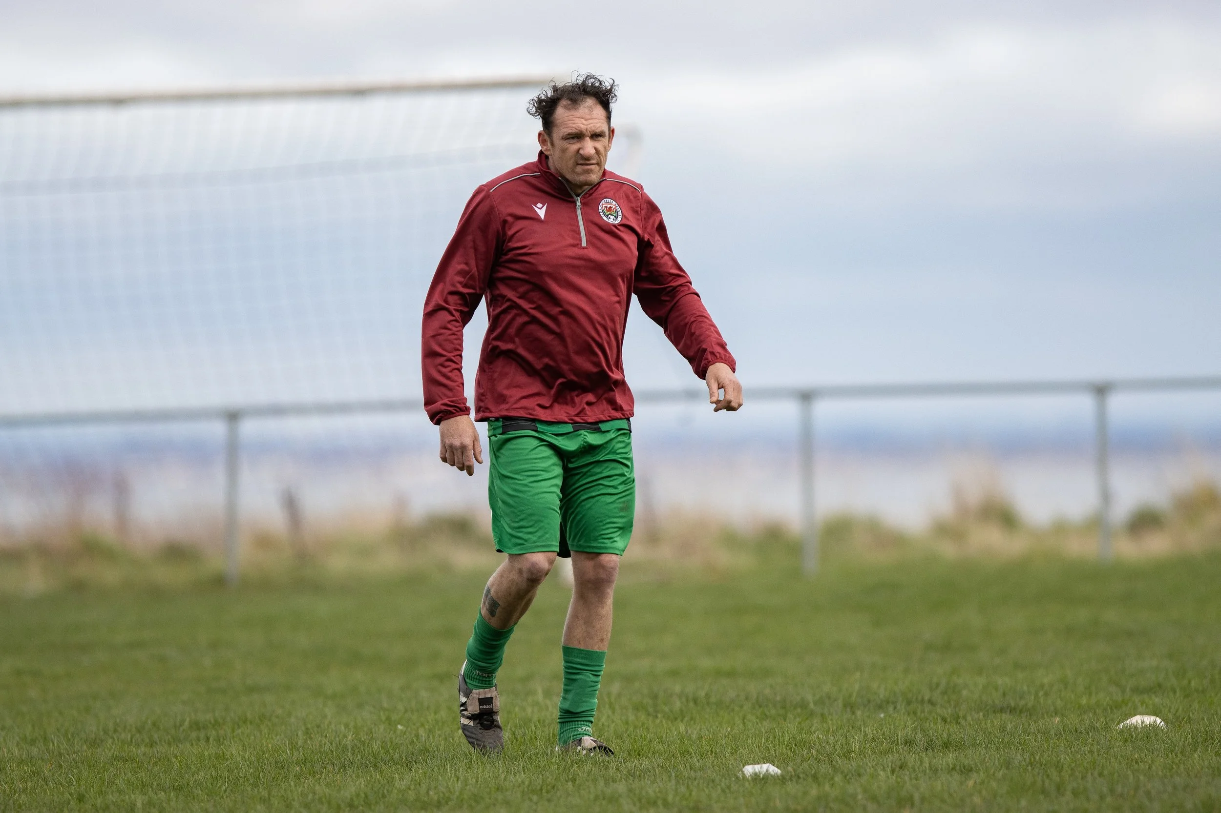 A man in a red sports jacket and green shorts walking on a soccer field with a goal net in the background.