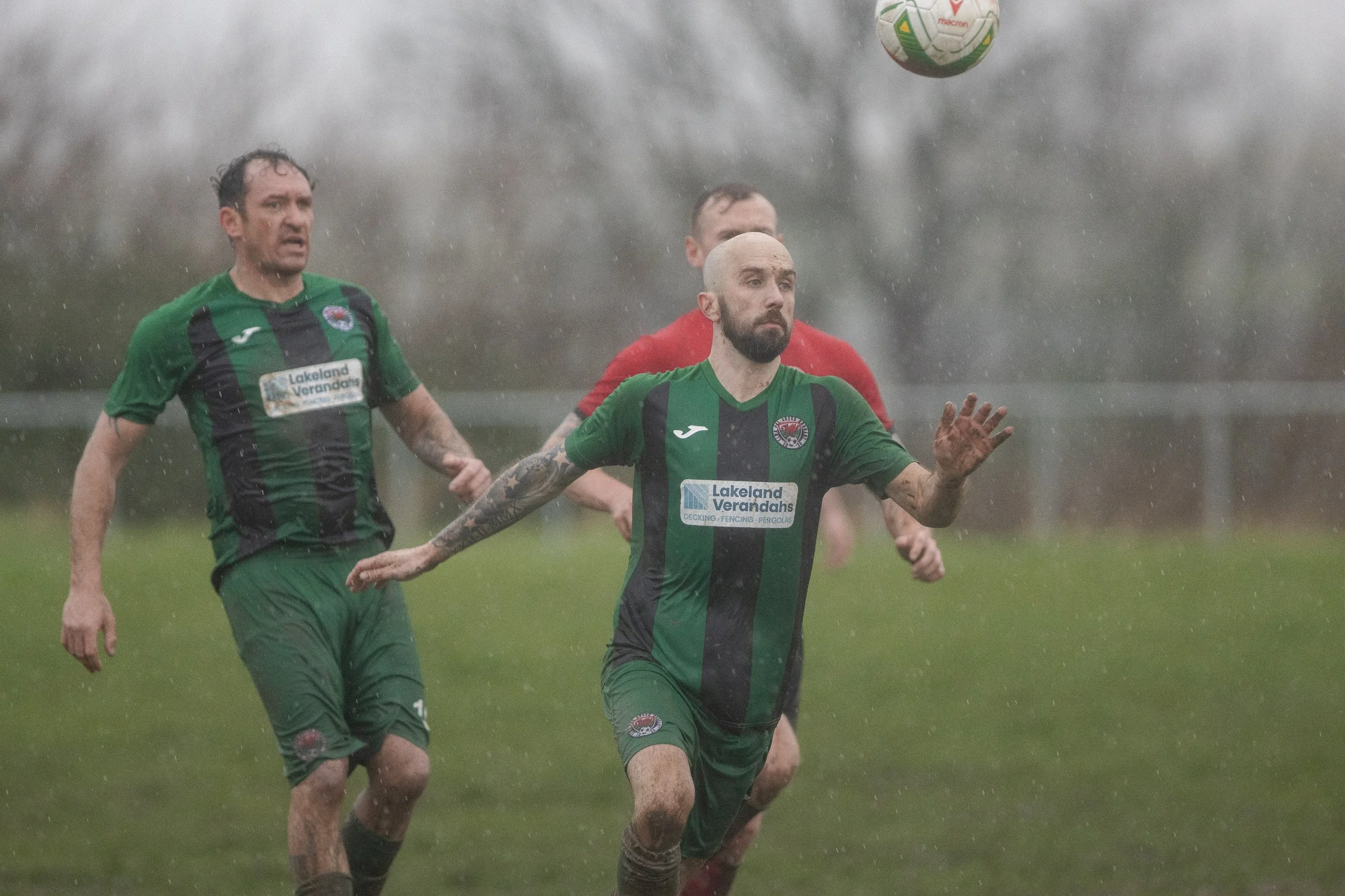Soccer players in green and red jerseys playing in a rainy outdoor field, chasing a soccer ball.