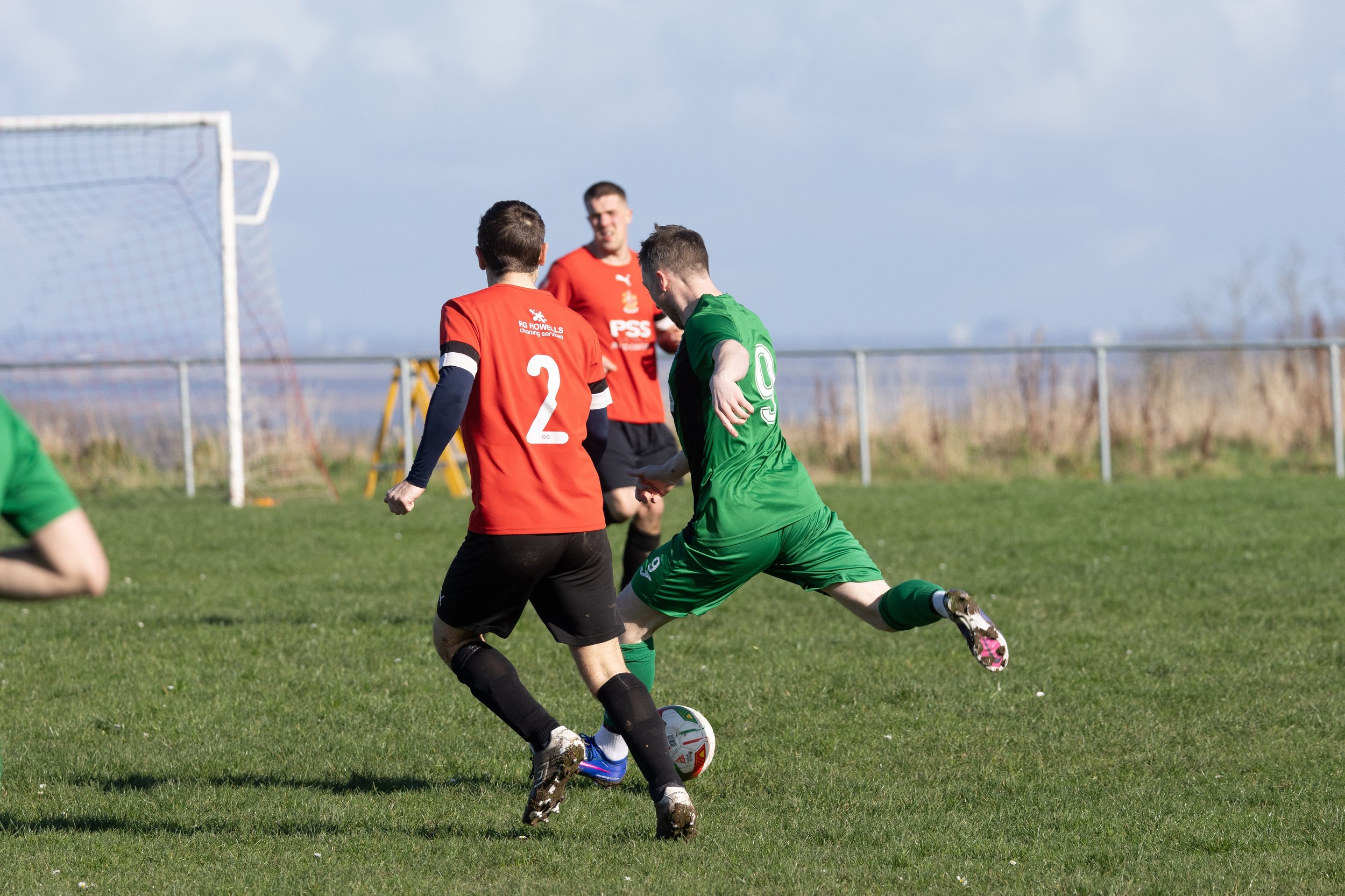 Soccer players in green and red jerseys competing for the ball on a grassy field, with goalpost and fence in the background.