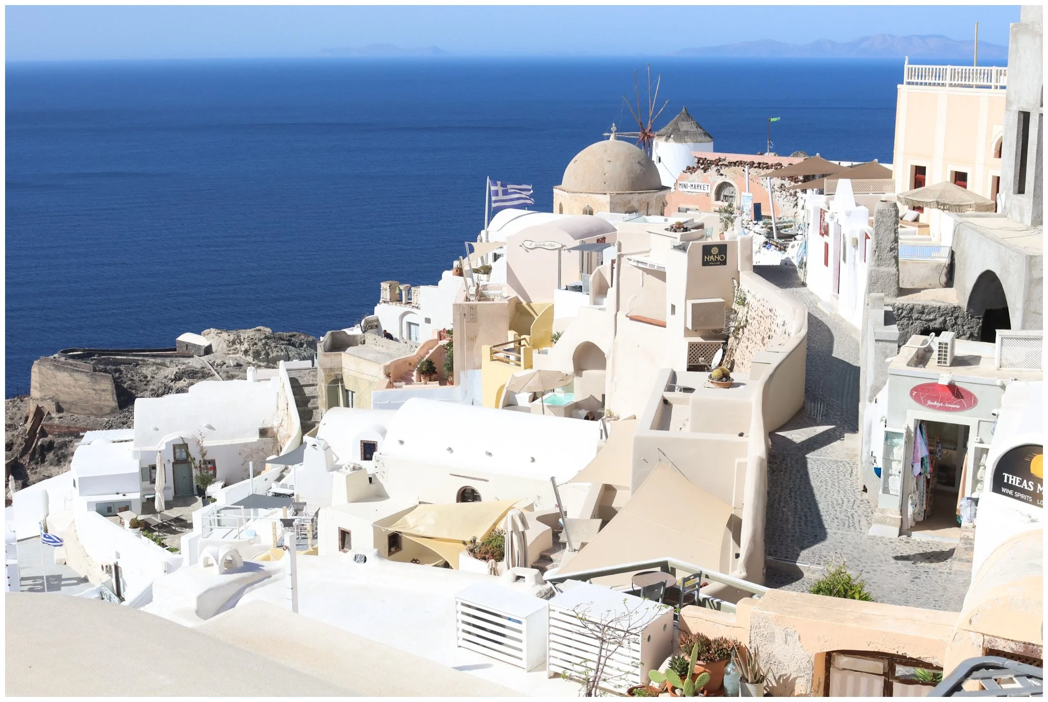 Scenic view of white buildings with flat roofs and awnings on a hillside overlooking the deep blue sea in Santorini, Greece, with clear sky and distant mountains.