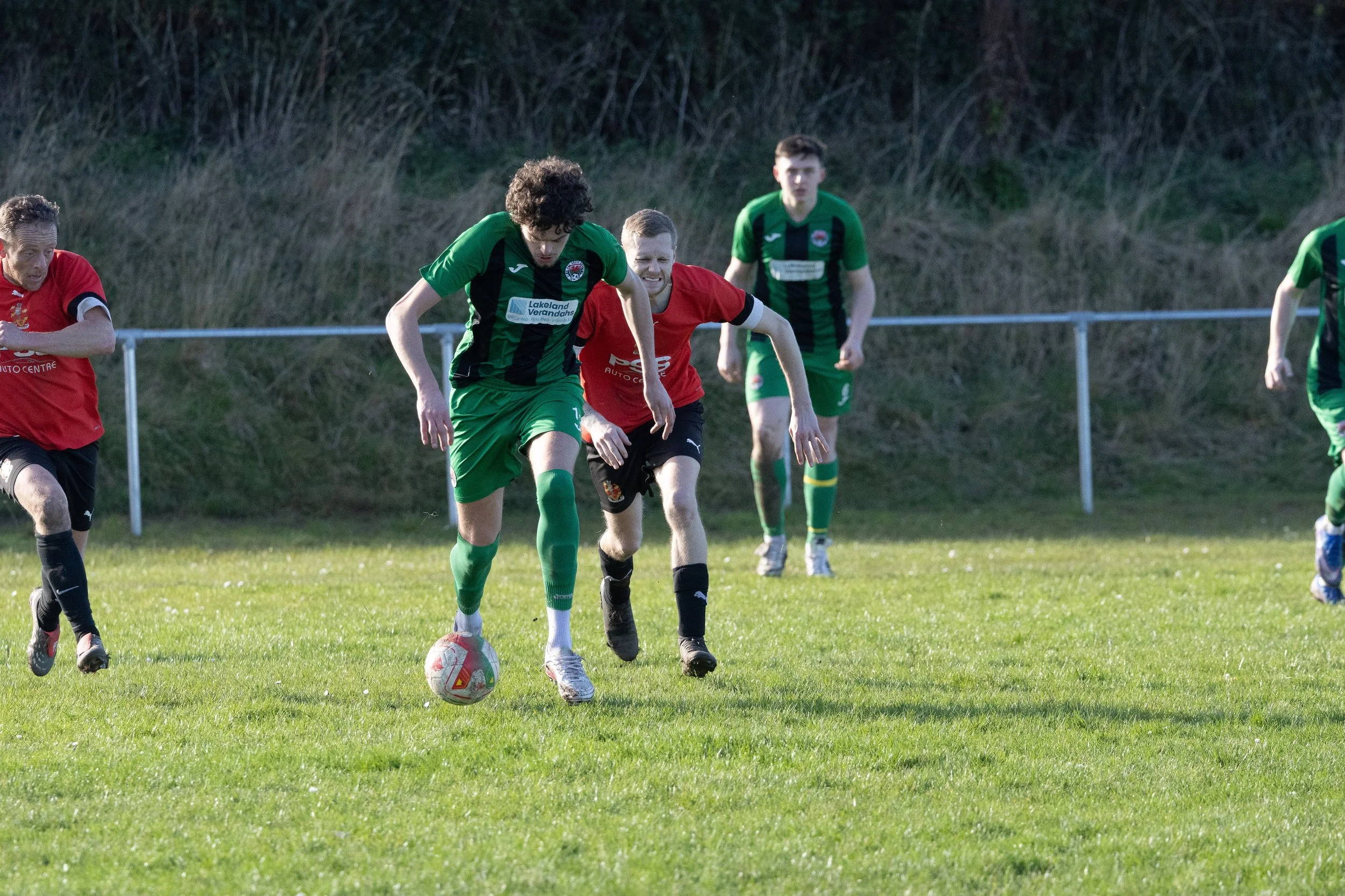 Soccer players in green and red uniforms competing for the ball on a grassy field.