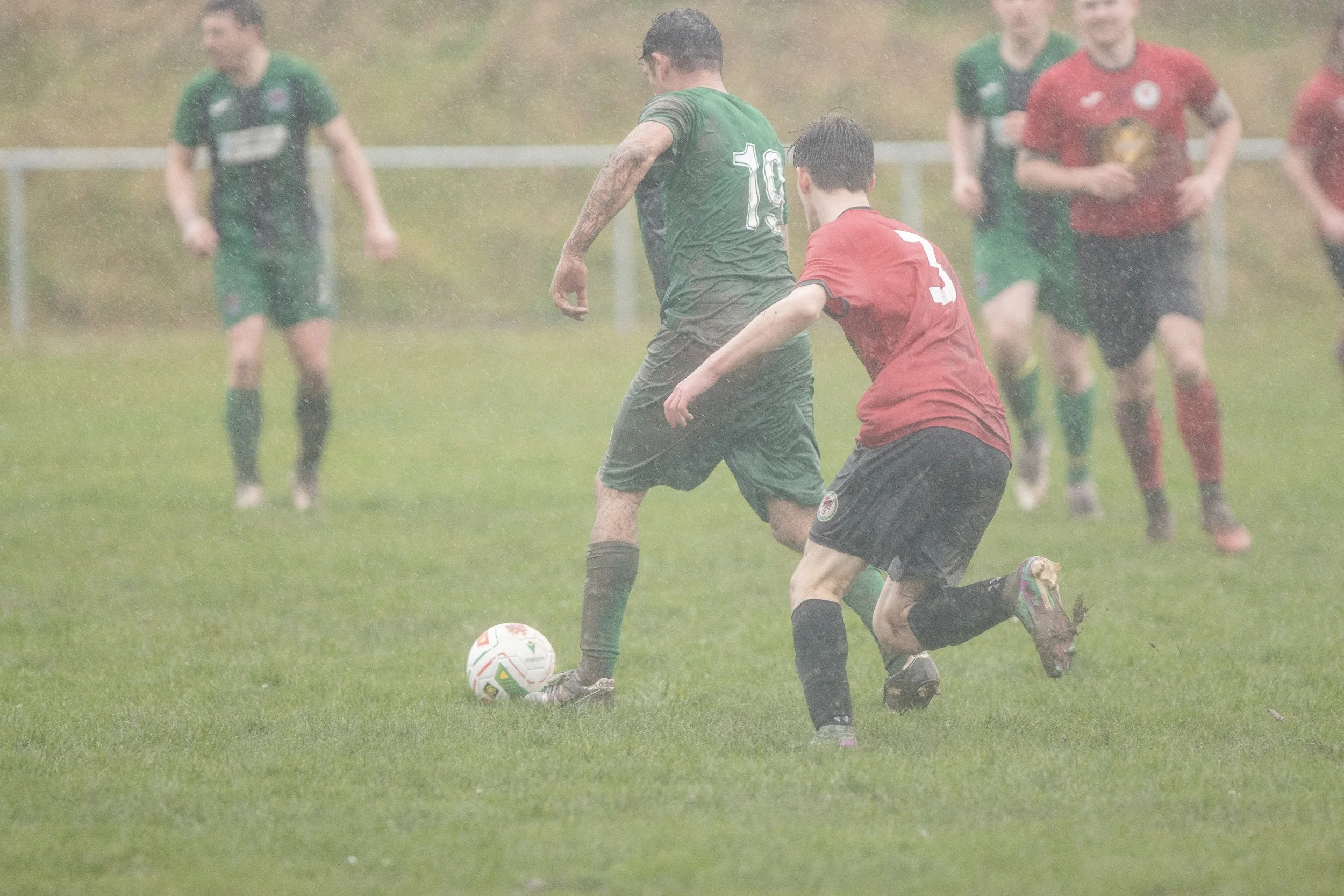 Two soccer players, one in a green jersey with the number 19 and the other in a red jersey with the number 3, compete for the ball in the rain on a grassy field, with four other players blurred in the background.