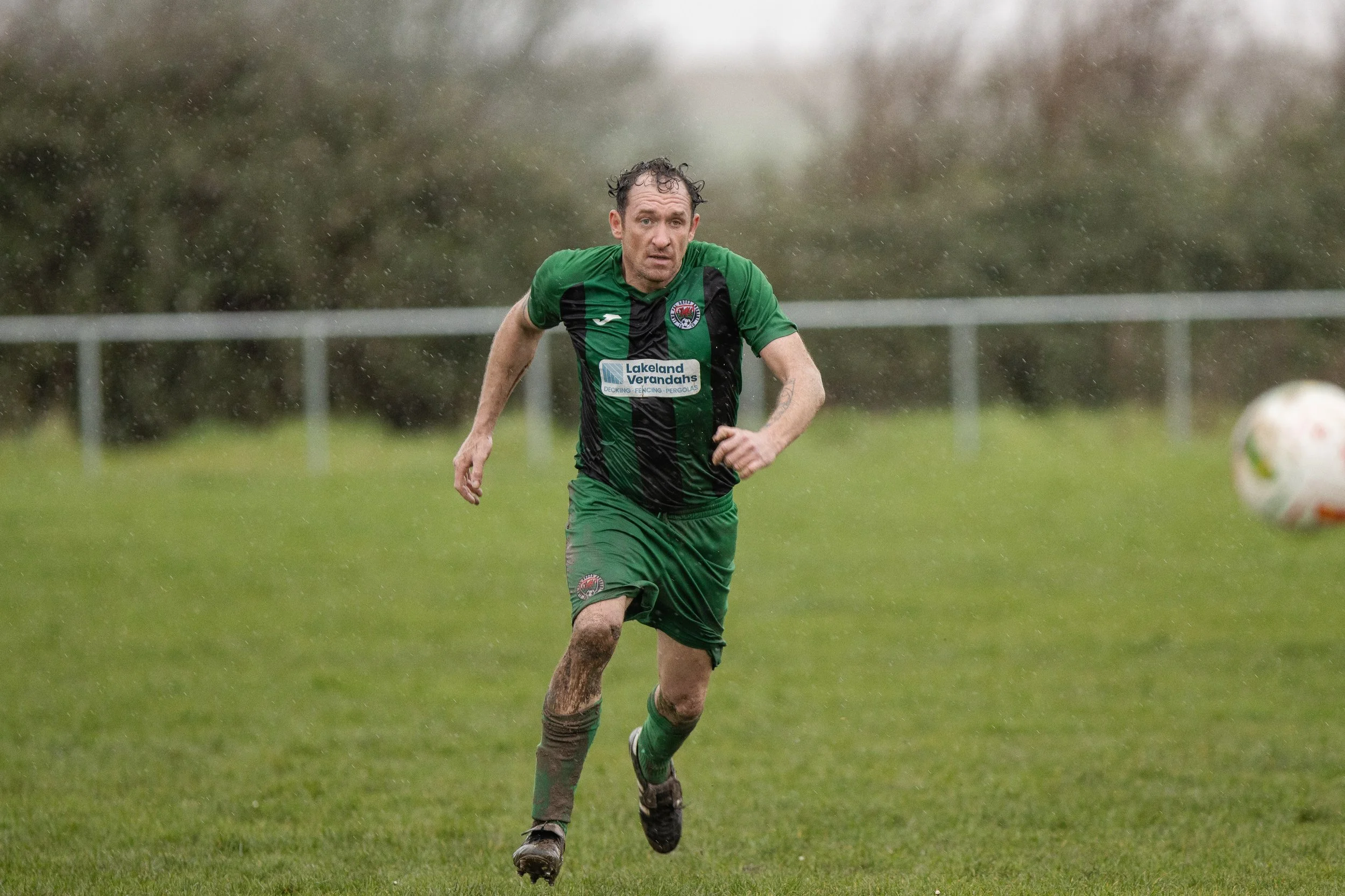 A male soccer player running on a rain-soaked field during a game, wearing a green and black uniform, with a focused expression as he pursues the ball.