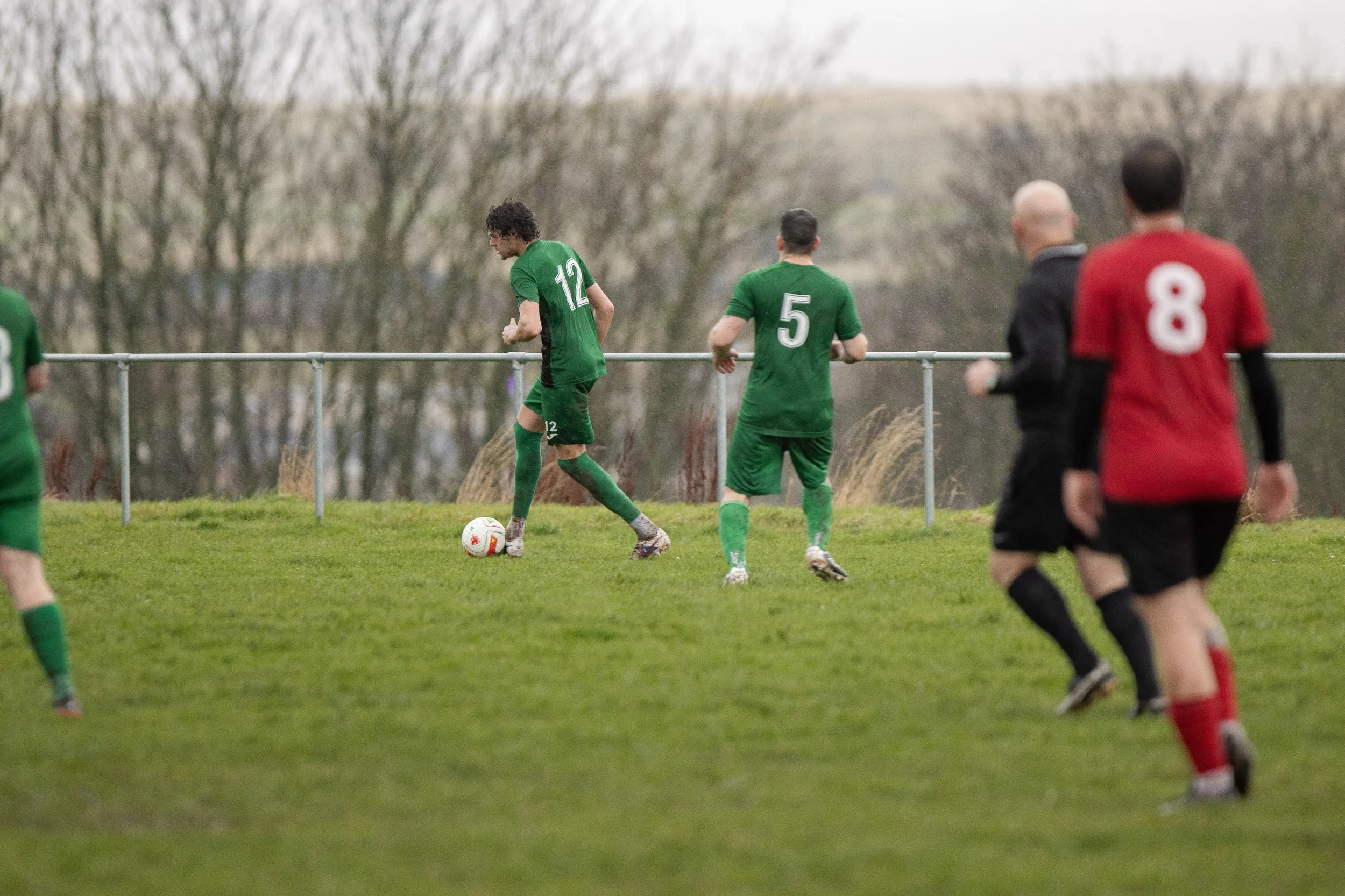 Soccer match in progress with players in green and red uniforms on a grassy field, some players running and others preparing for the game.