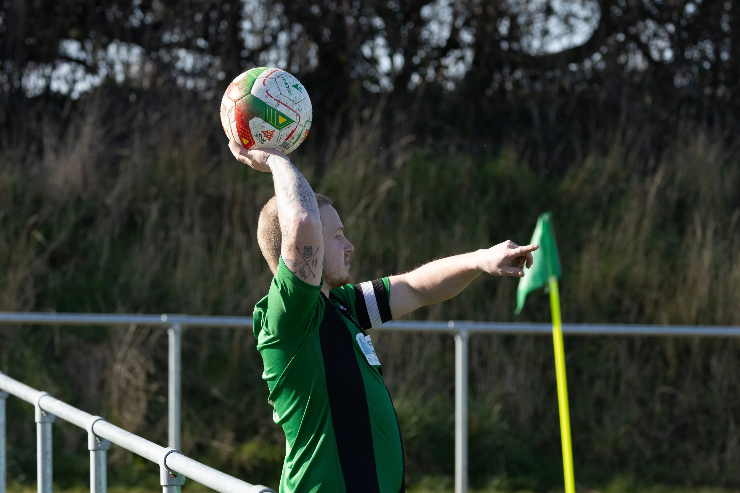 A soccer player in a green jersey holding a ball in his right hand and pointing with his left hand on the field.