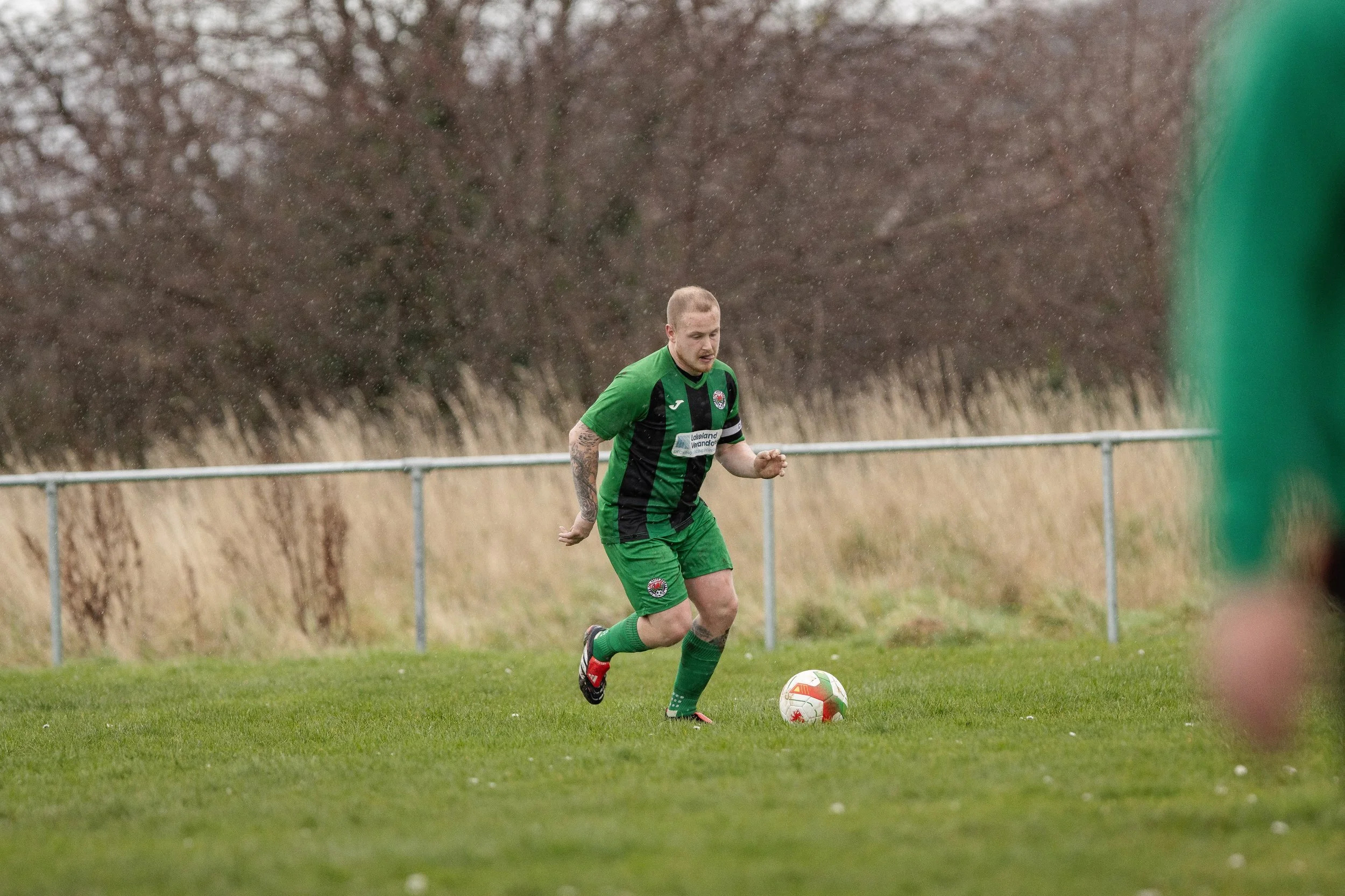 A soccer player in a green and black uniform is on the field, preparing to kick a soccer ball. The background shows a grassy area with trees and a metal fence.