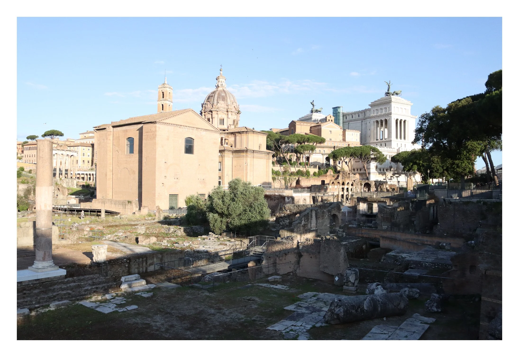 Ancient Roman ruins in Rome with classical buildings and trees in the background under a blue sky.