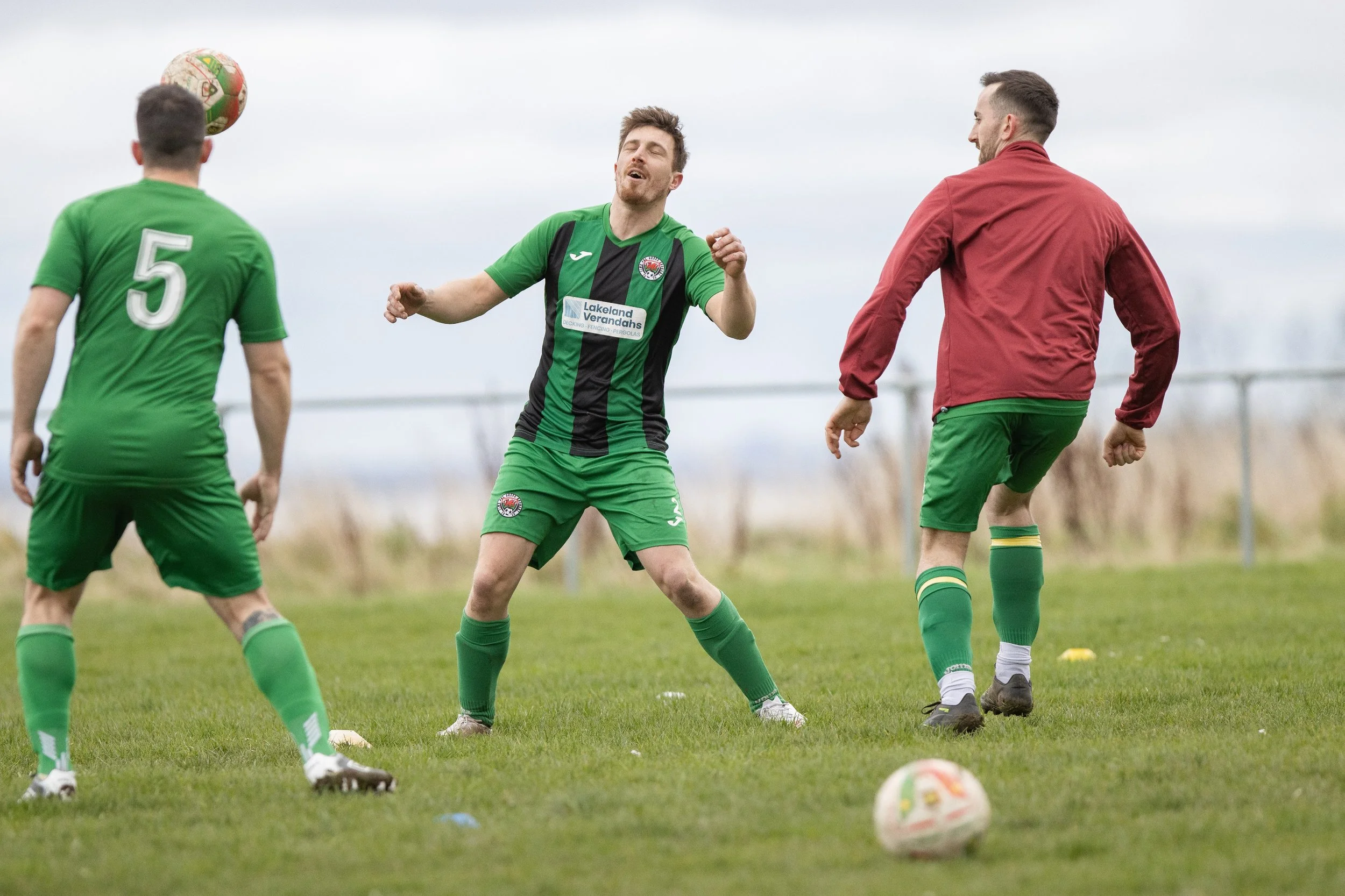 Three men playing soccer on a grass field, one wearing a red jacket, and the other two in green jerseys, with one of the green jersey players about to kick the ball.