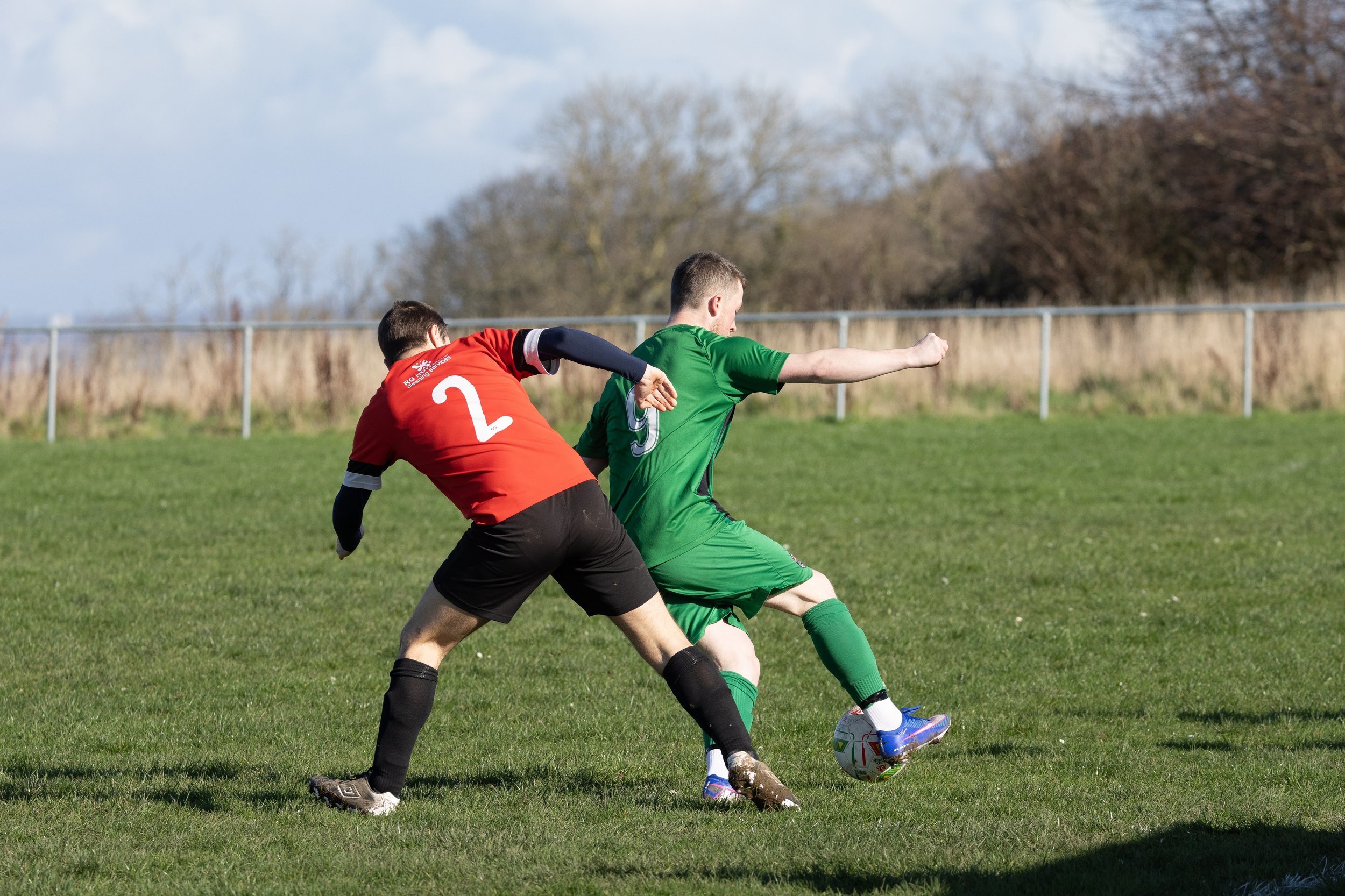 Two soccer players competing for the ball on a grassy field, one in a red uniform with the number 2 and the other in a green uniform with the number 9, during daytime.
