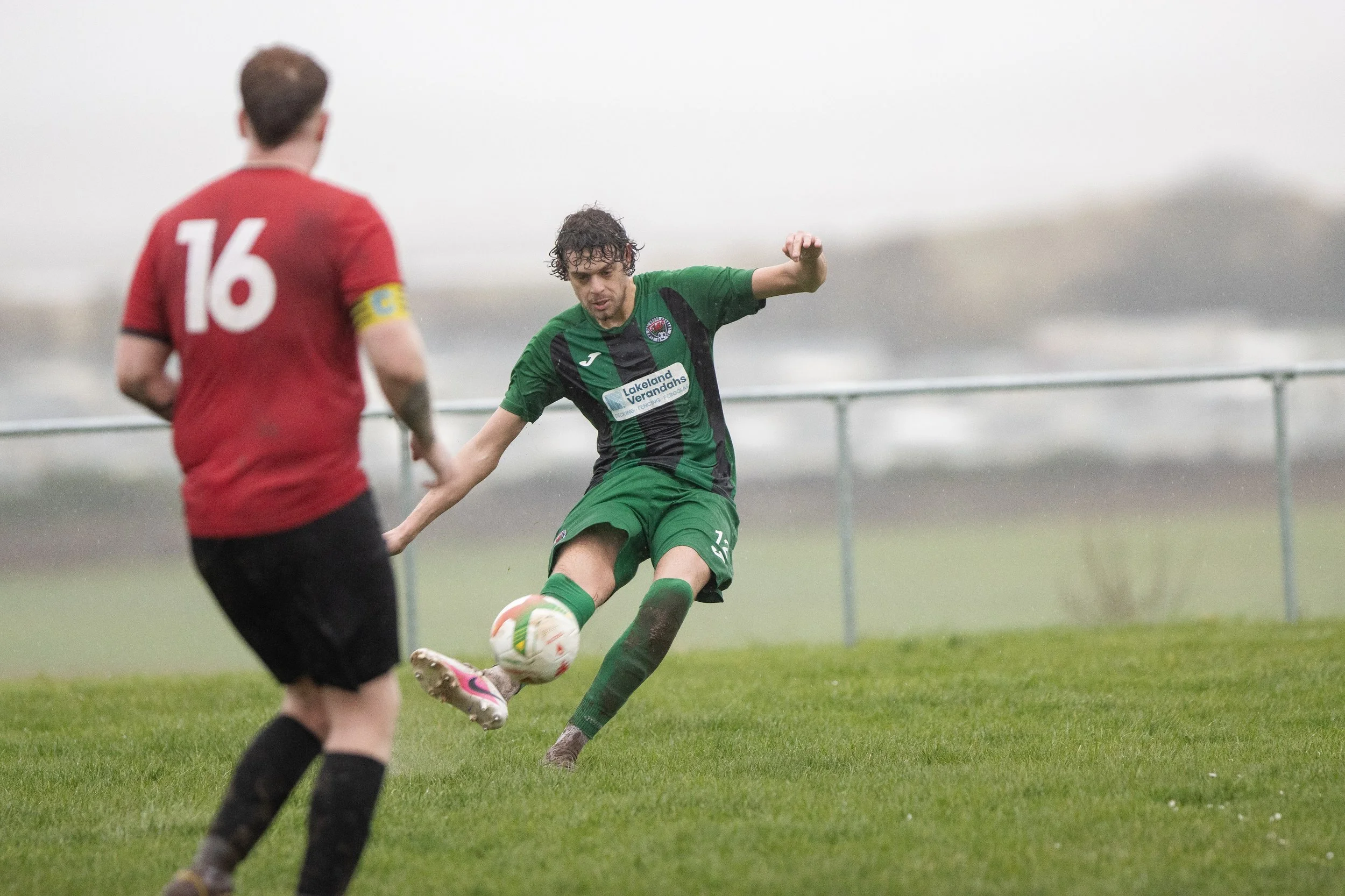A soccer player in a green uniform kicks a soccer ball while a player in a red jersey watches during a game on a grassy field in rainy weather.