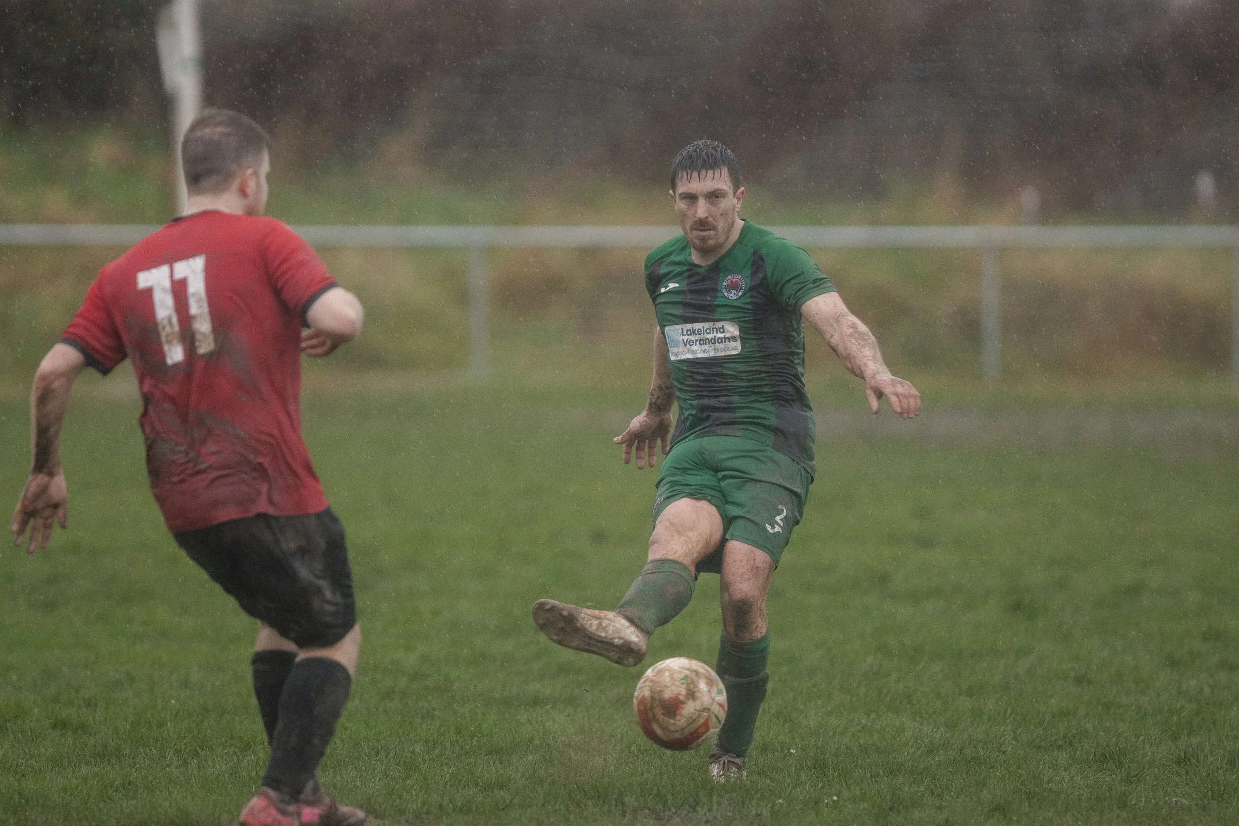 A soccer match in the rain, featuring two players: one in a red jersey with the number 11 on the back, and the other in a green kit. The player in green is kicking the ball, muddy and soaked, on a grass field with a fence in the background.