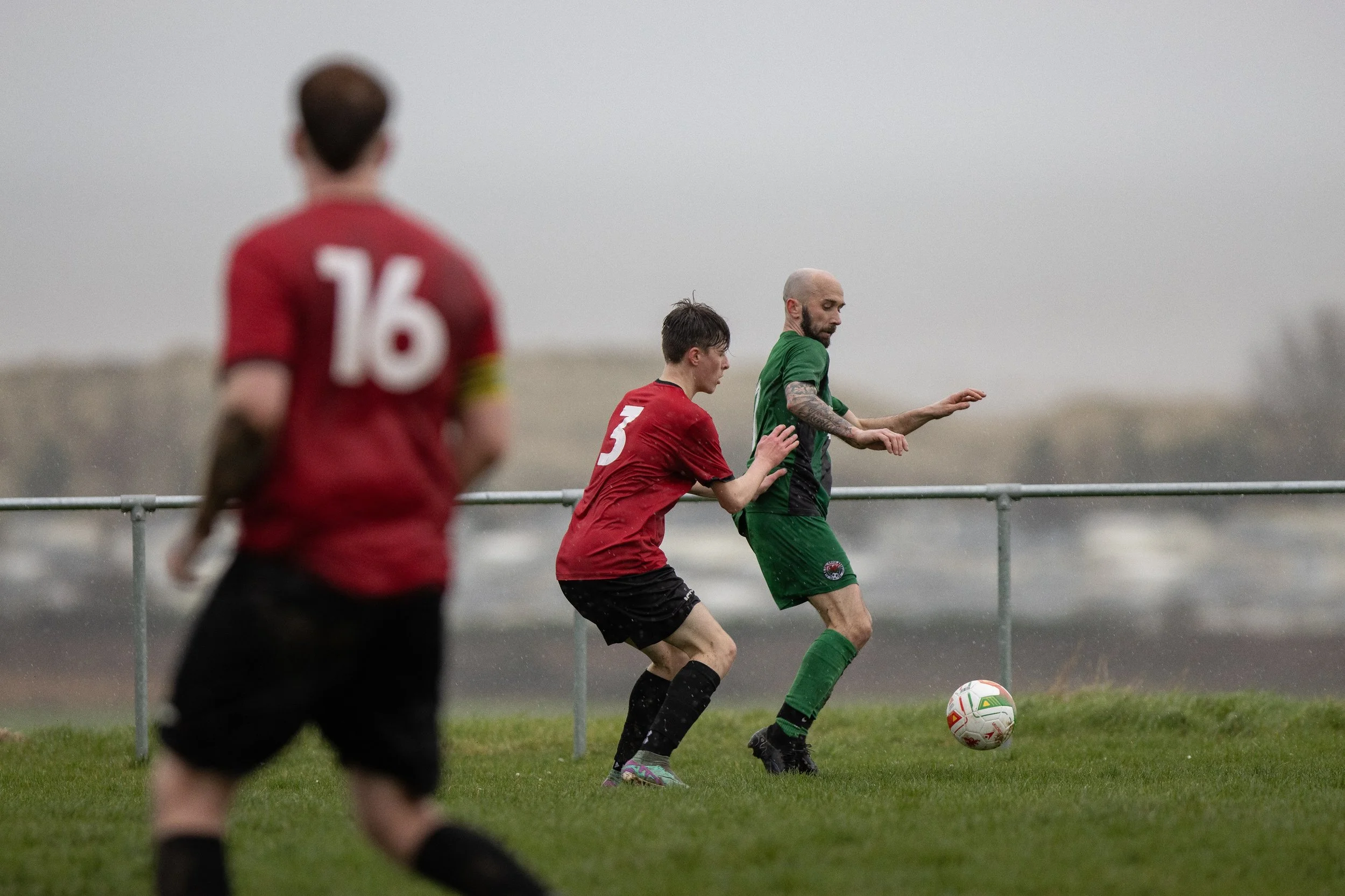 Three soccer players in red and green uniforms playing soccer outdoors on a grassy field on a rainy day, with a cloudy sky and blurred landscape in the background.