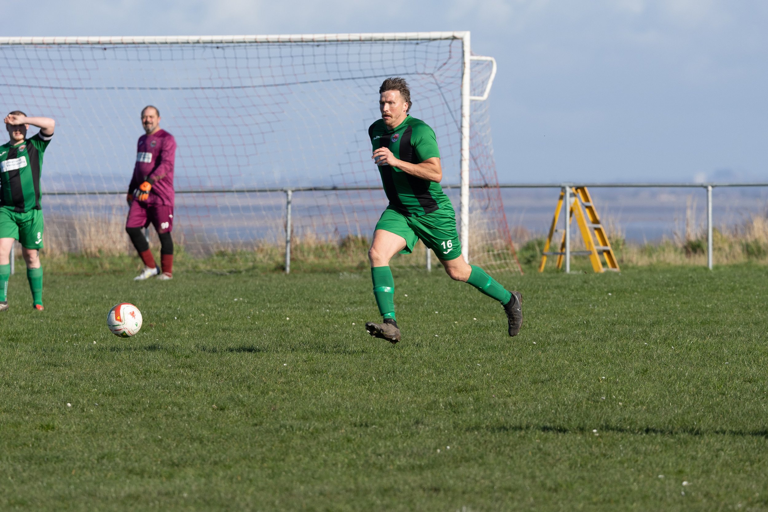 A soccer player in a green and black uniform running on the field with a soccer ball nearby. There are other players and a goalkeeper in the background.
