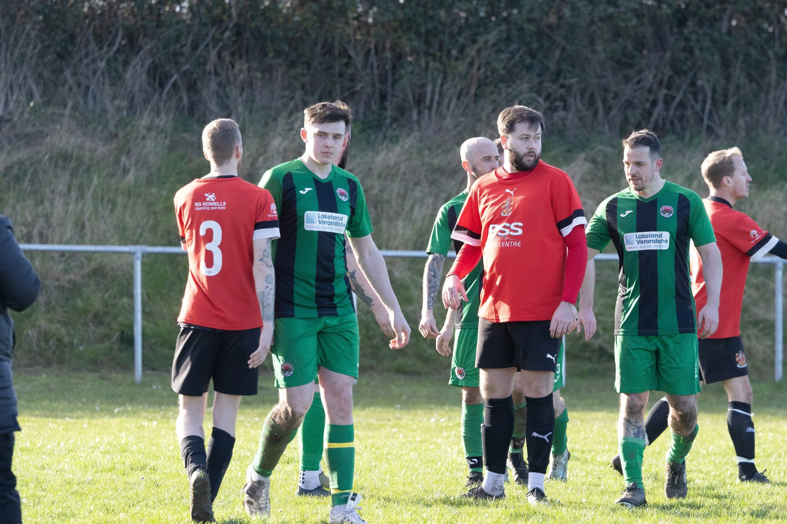 Soccer players in green and red uniforms on the field after a match, standing on grass with a natural background.