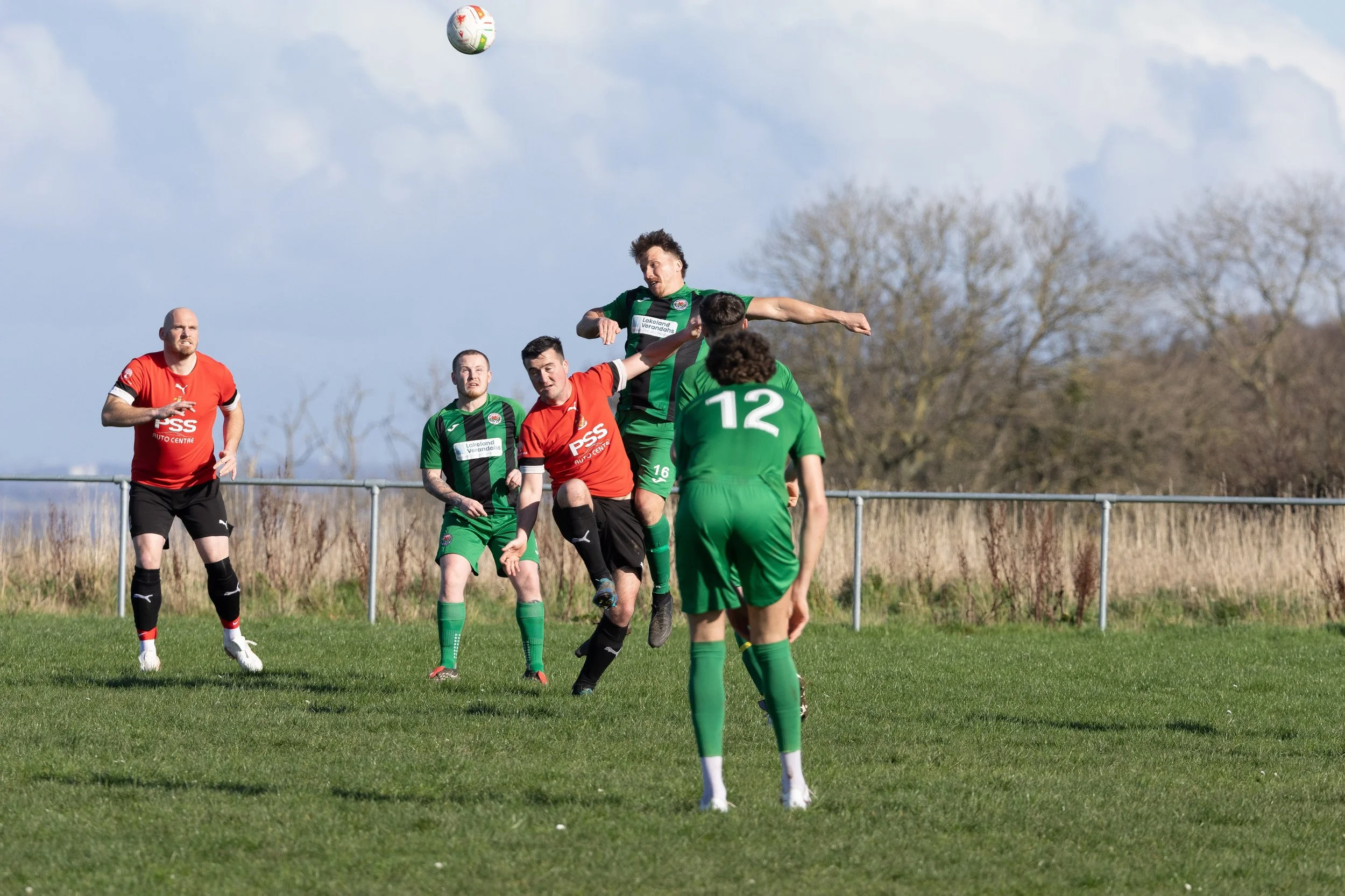 Soccer players in green and red uniforms competing for a ball in mid-air on a grassy field, with trees and a fence in the background.