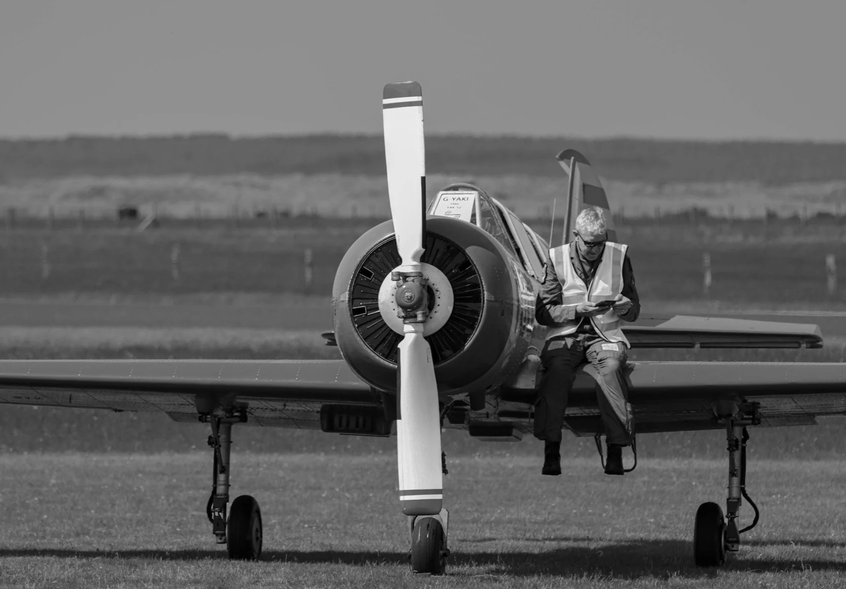 A man in a safety vest sitting on the front of a small aircraft, reading a book or tablet, with a rural landscape in the background.