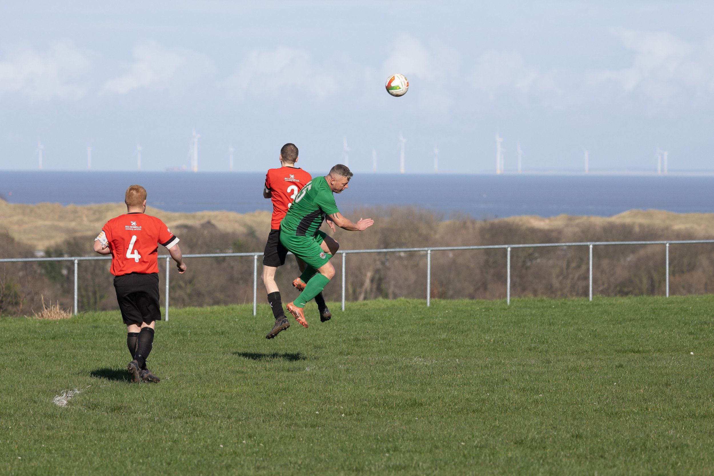 Four soccer players on a grass field near the coast, with wind turbines in the background. Two players in red jerseys are facing the camera, while a player in green and black is jumping to head the ball. The ball is in mid-air above the green player.