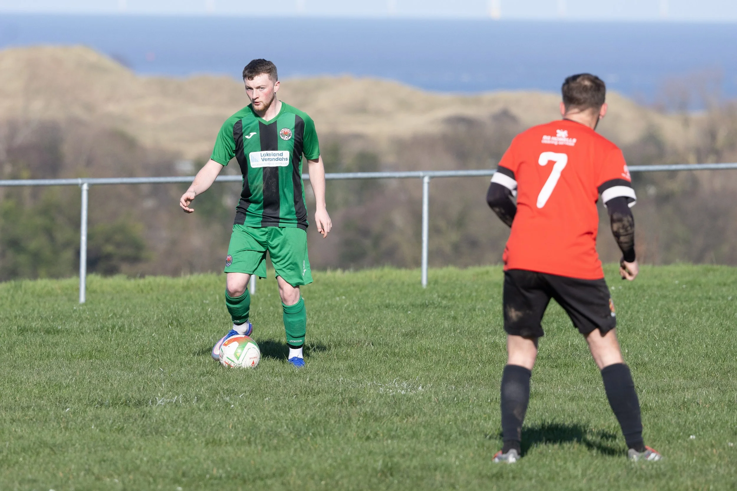 Two soccer players on a grassy field, one in green and black uniform preparing to kick the ball, the other in red and black uniform with the number 7 on the back, facing away.