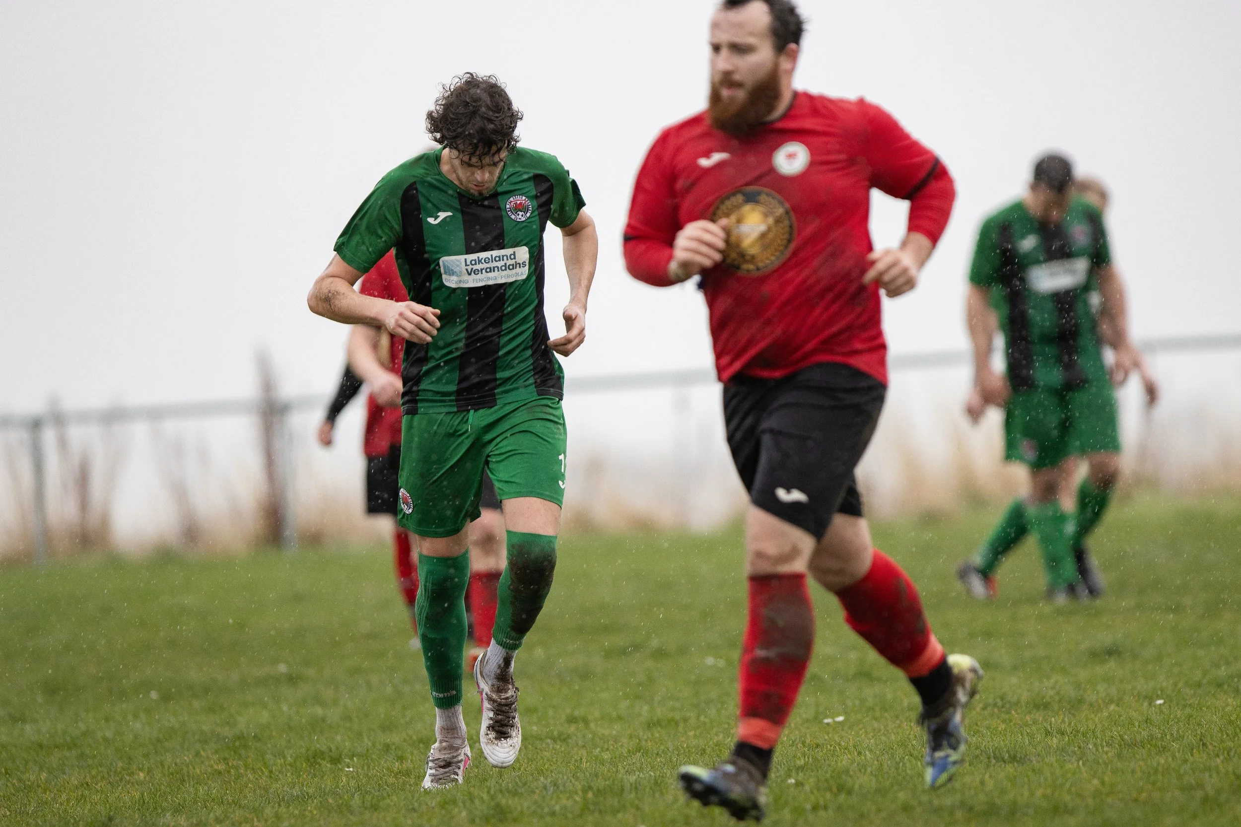 Soccer players running on a wet field during a match in rainy weather. The player in the foreground is wearing a red jersey and black shorts, while the player behind is wearing a green and black striped uniform with green shorts.