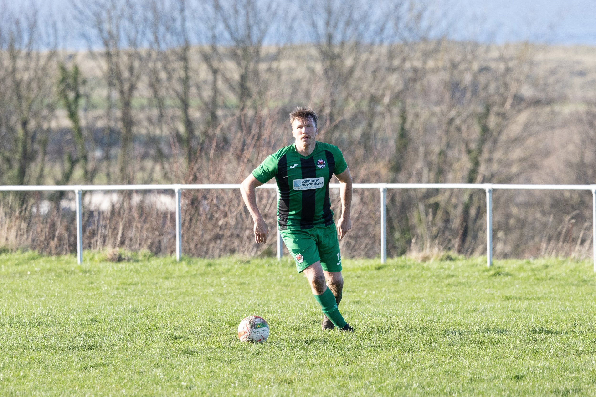A soccer player in a green and black jersey preparing to kick a soccer ball on a grassy field, with a background of trees and a white fence.