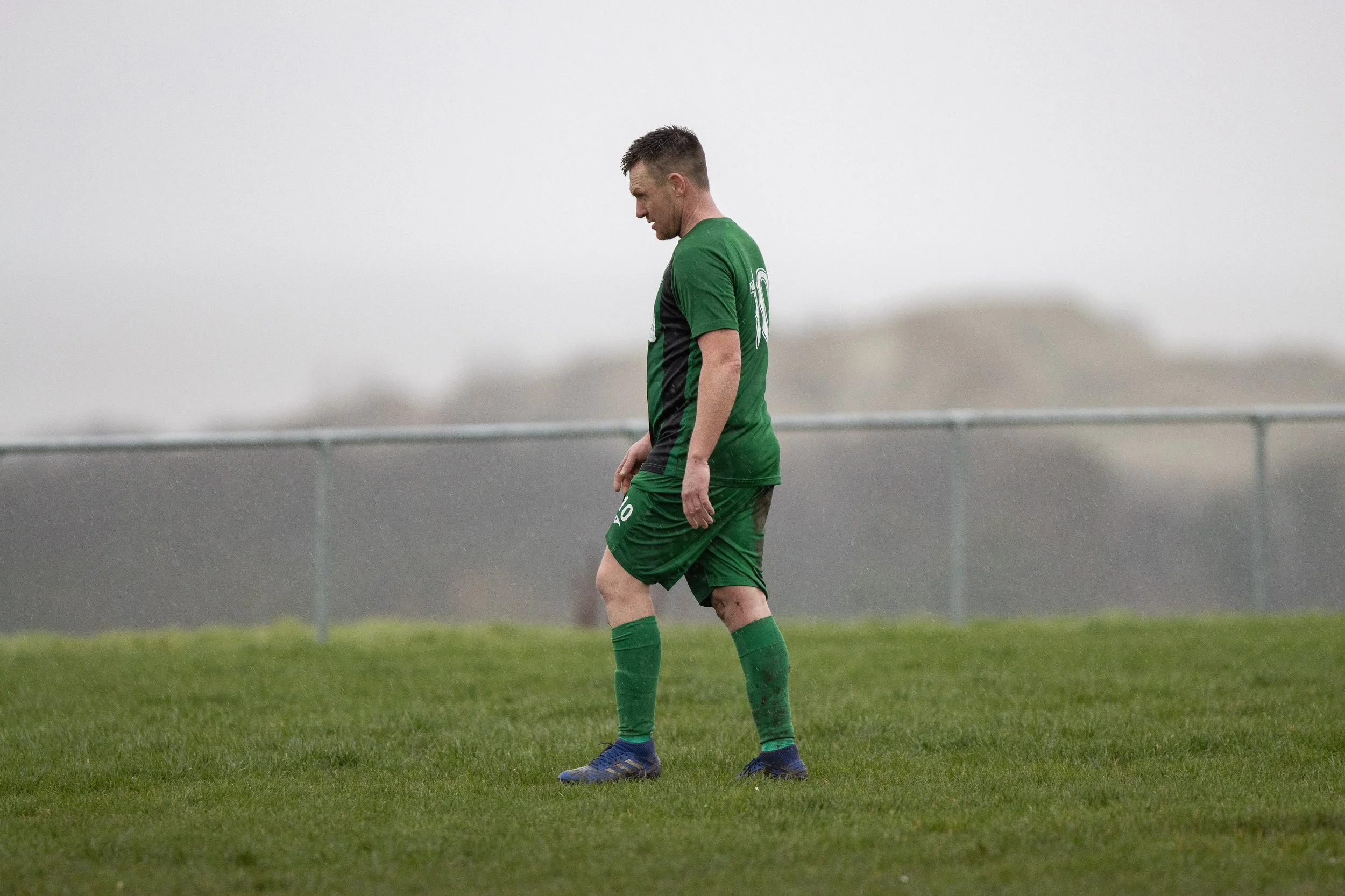 A soccer player in a green uniform standing on a grass field on a rainy day.