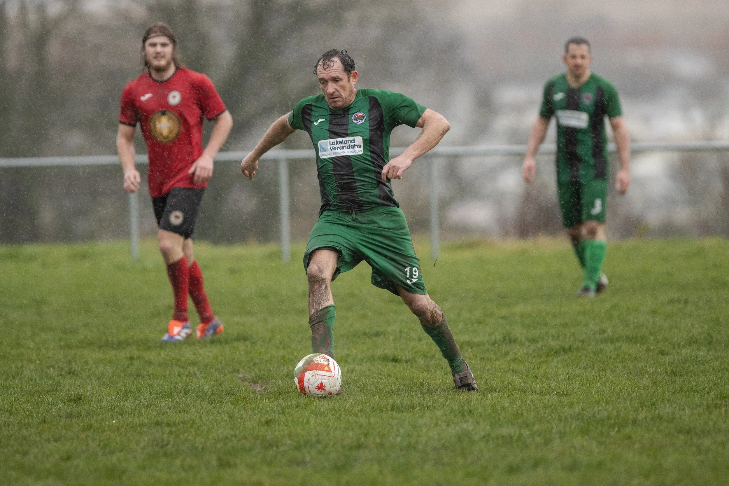 A soccer player in a green and black uniform kicking a ball on a rainy field, with two other players in red and green uniforms in the background.