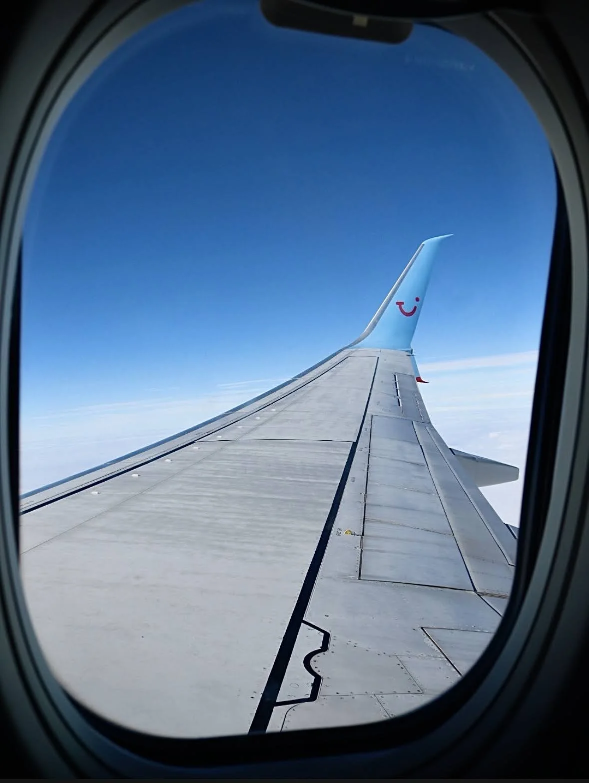 View of airplane wing through aircraft window with blue sky and clouds in the background.