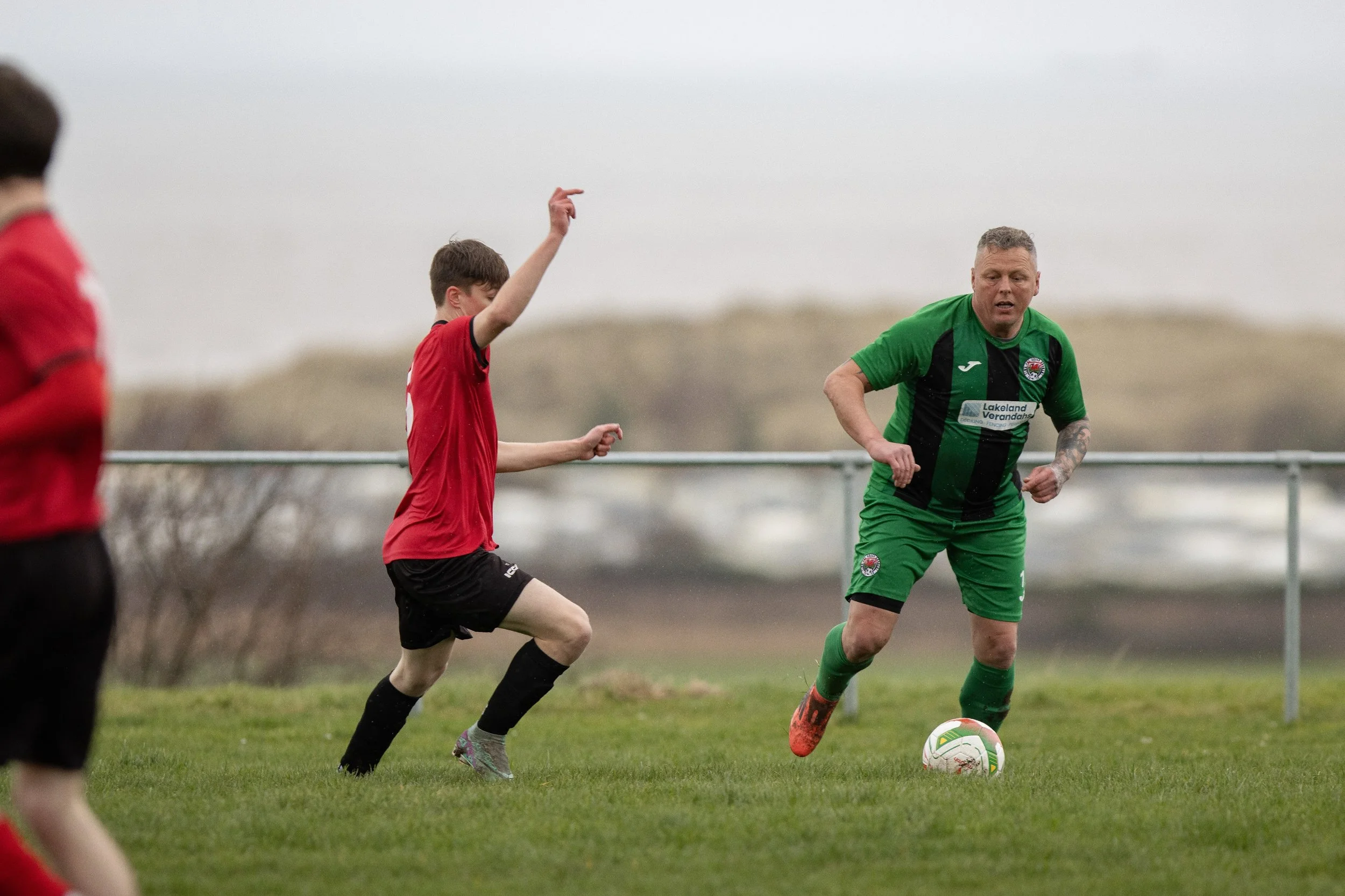 A man in a green and black soccer uniform dribbling a soccer ball on a grassy field while young players in red jerseys observe, with cloudy sky and distant trees in the background.
